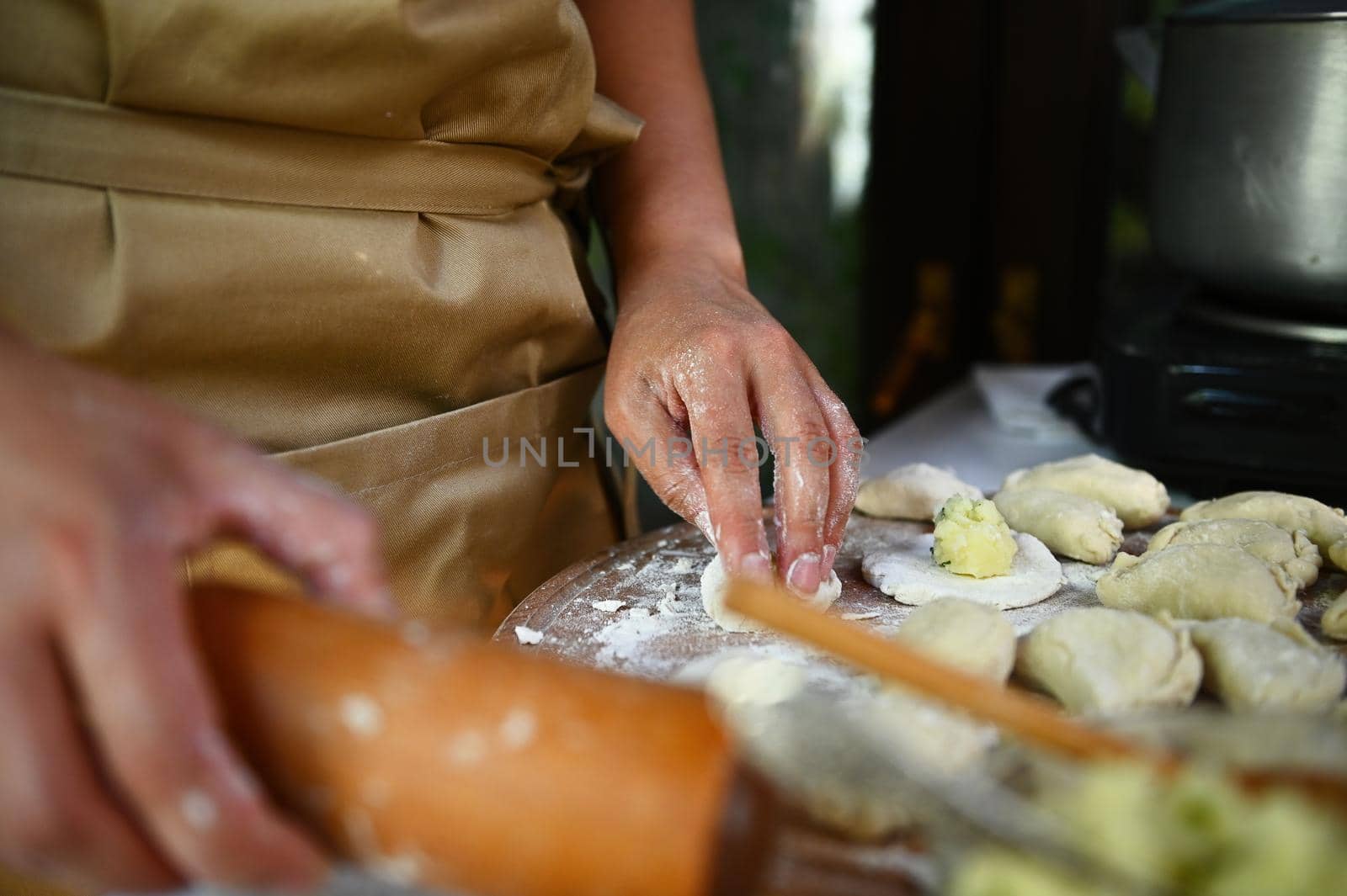 Selective focus on housewife in beige chef's apron, rolling out the dough in a rustic kitchen, making homemade dumplings, according to traditional Ukrainian recipe. Vareniki. Pelmeni. Italian Ravioli