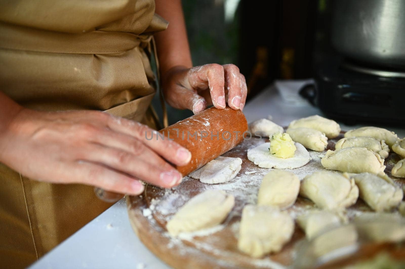 Selective focus. Close-up: Chef in a beige apron, using a rolling pin, rolls out dough on a wooden board, prepares homemade dumplings or Ukrainian national dish Varenyky. Cooking class. Baking concept