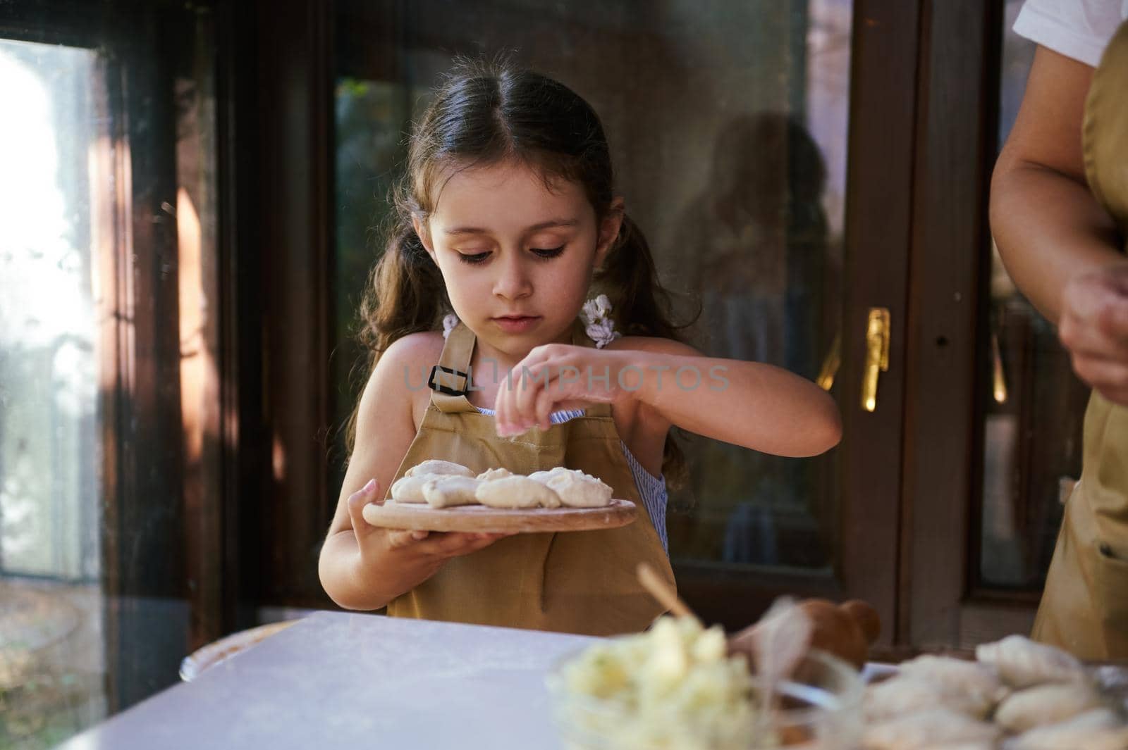 Beautiful little girl wearing chef's apron, standing at kitchen table and holding a wooden board with homemade dumplings by artgf