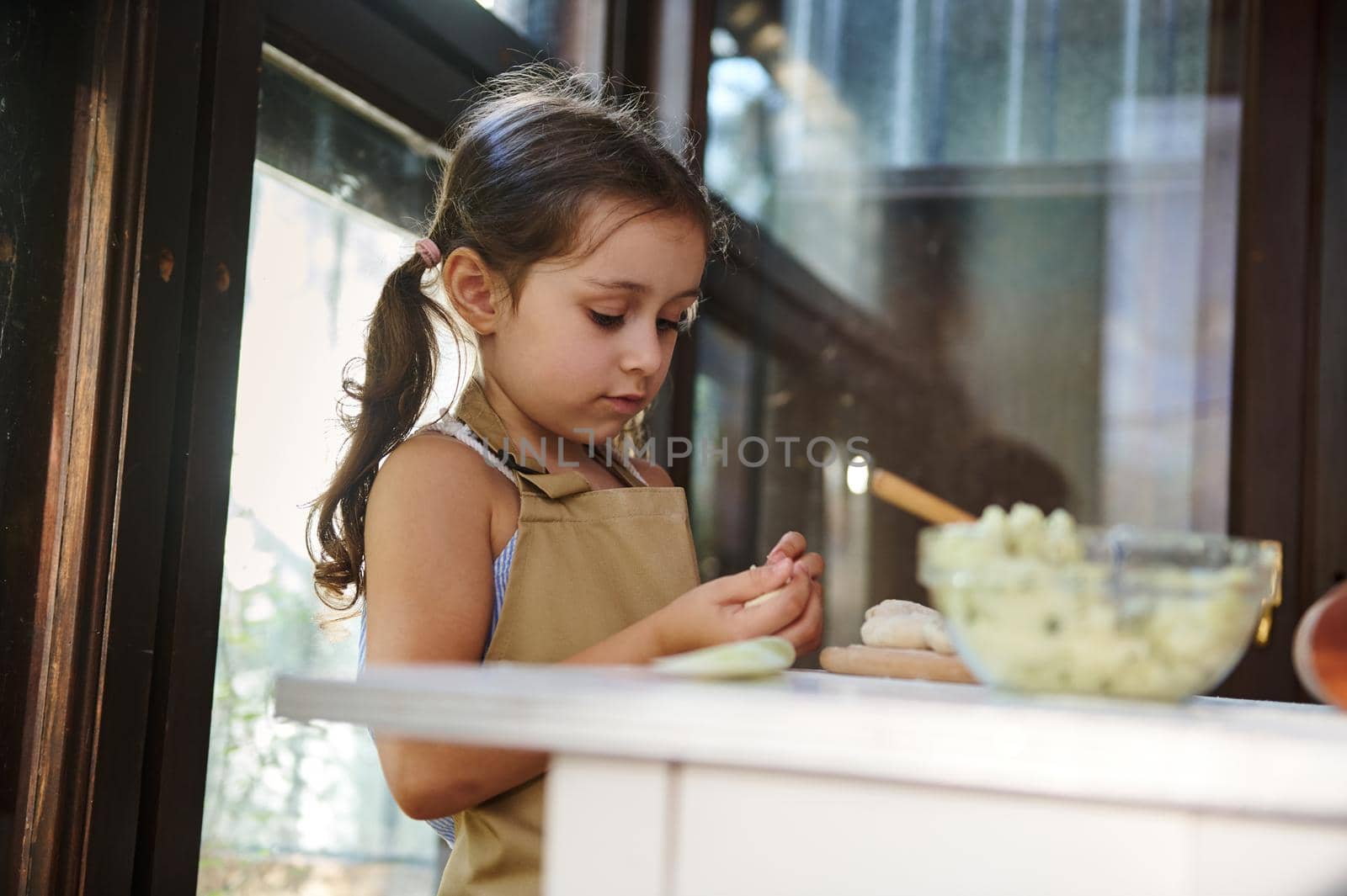 Close-up of a beautiful Caucasian child, a little cook, adorable baby girl wearing beige chef's apron hands-on dumplings in a country kitchen, helping her mother cooking Ukrainian varenyky for dinner