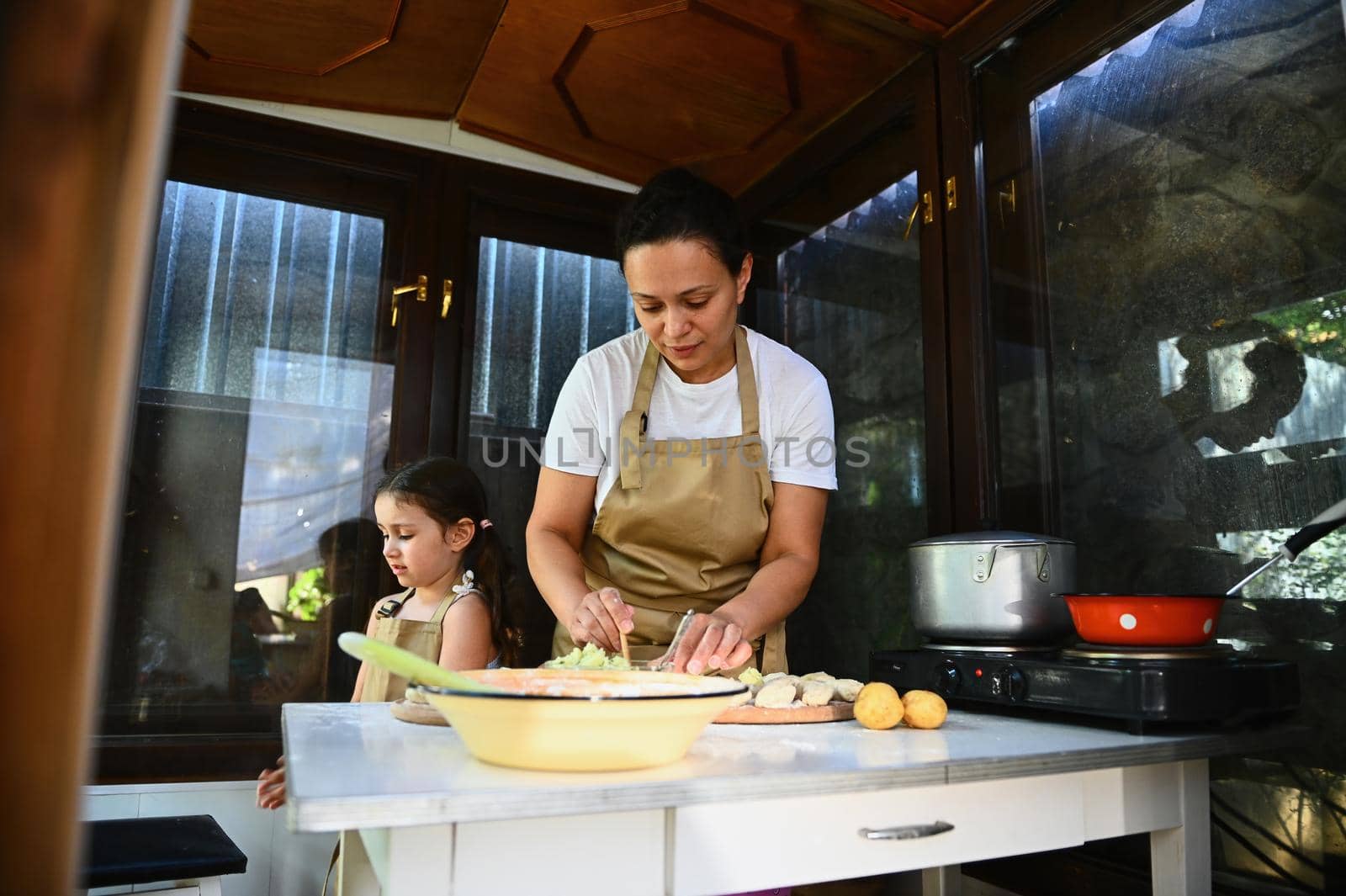 Loving mother cooking dumplings in the country kitchen. A housewife filling rolled dough with mashed potatoes, preparing a delicious lunch with her little daughter, in the village in the summer day