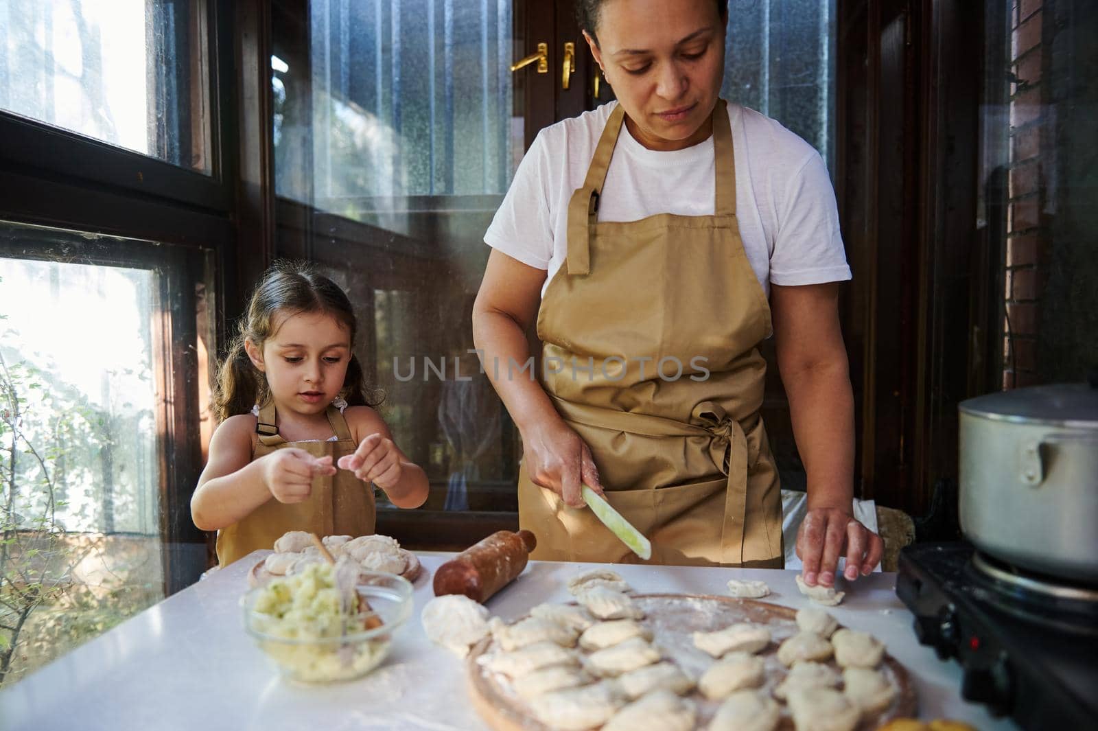 Loving mother and her adorable little daughter molding dumplings. Multi-ethnic pretty woman in a beige chef's apron, a housewife cooking Ukrainian vareniki in the rural summer kitchen. Baking concept