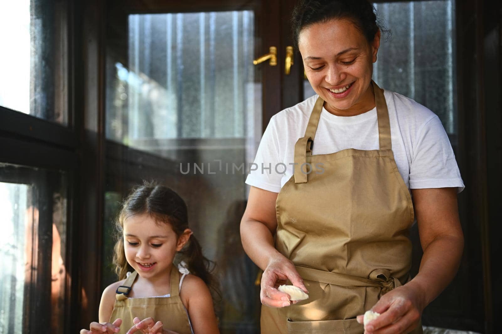 Pretty cheerful middle-aged multi-ethnic woman, a loving mom and her cute little daughter, both of them in beige chef's aprons, cooking dumplings, Ukrainian varenyky for dinner in the country kitchen