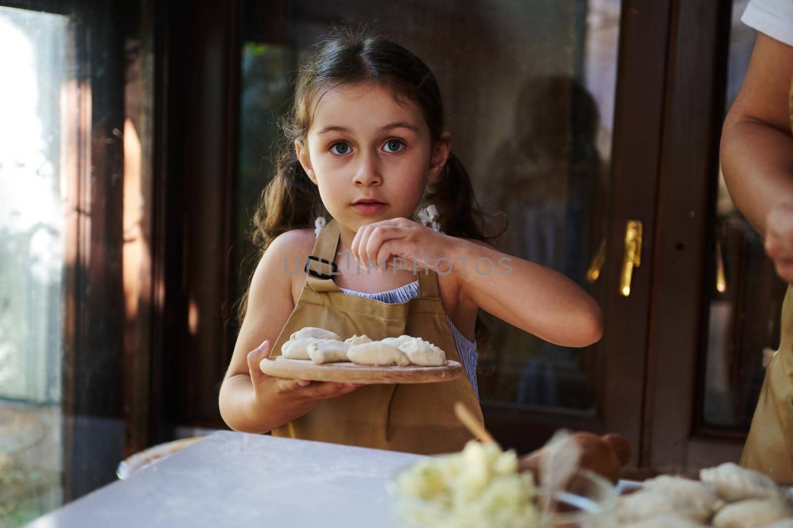 Beautiful Caucasian child, little girl wearing beige chef's apron stands next to her mother and sprinkling flour on a wooden board with homemade dumplings. Kid learning culinary. Baking master class