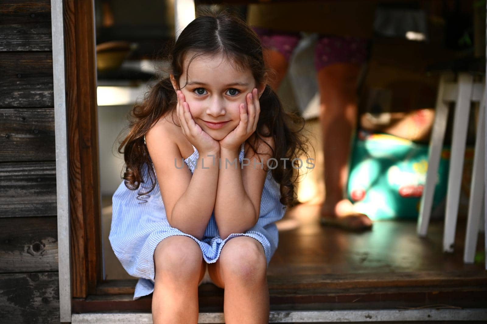 Confident portrait of an adorable 5 years old child, cute little girl, in a blue sundress, sitting on the doorstep of a country kitchen and looking at camera. Beautiful people. Children. Childhood