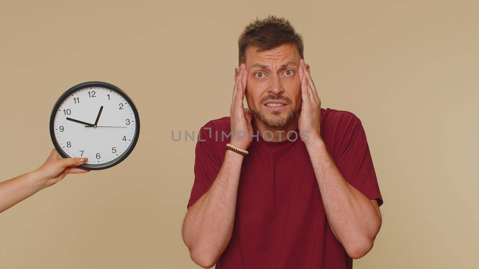 Confused young man in red t-shirt with anxiety checking time on clock, running late to work, being in delay, deadline. Adult stylish male guy looking at hour, minutes, worrying to be punctual, indoors