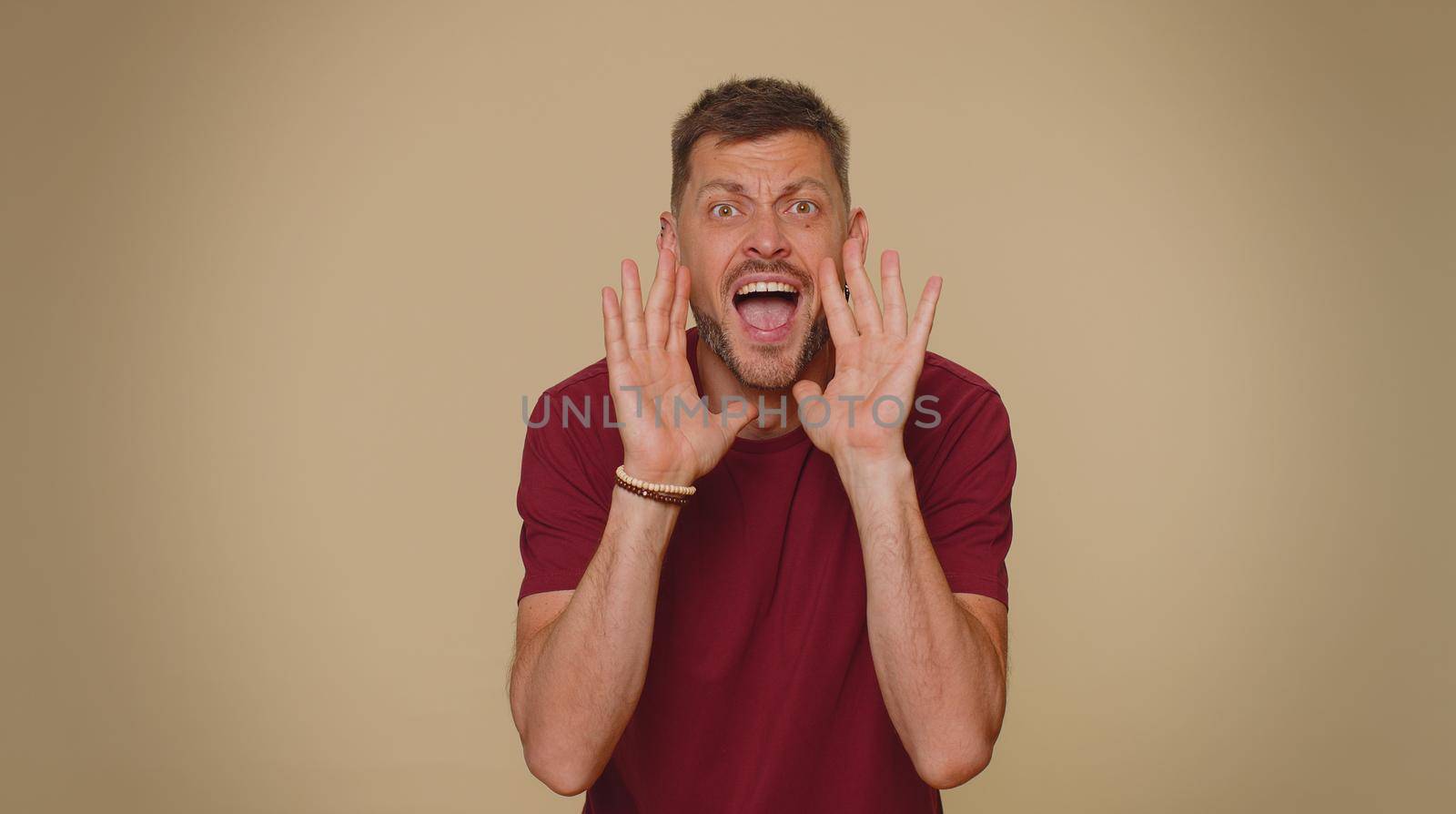 Expressive bearded handsome young man holding hands near mouth loudly screaming, shouting, yelling, announces discounts sale Hurry up, welcomes someone hello. Adult stylish guy on beige background