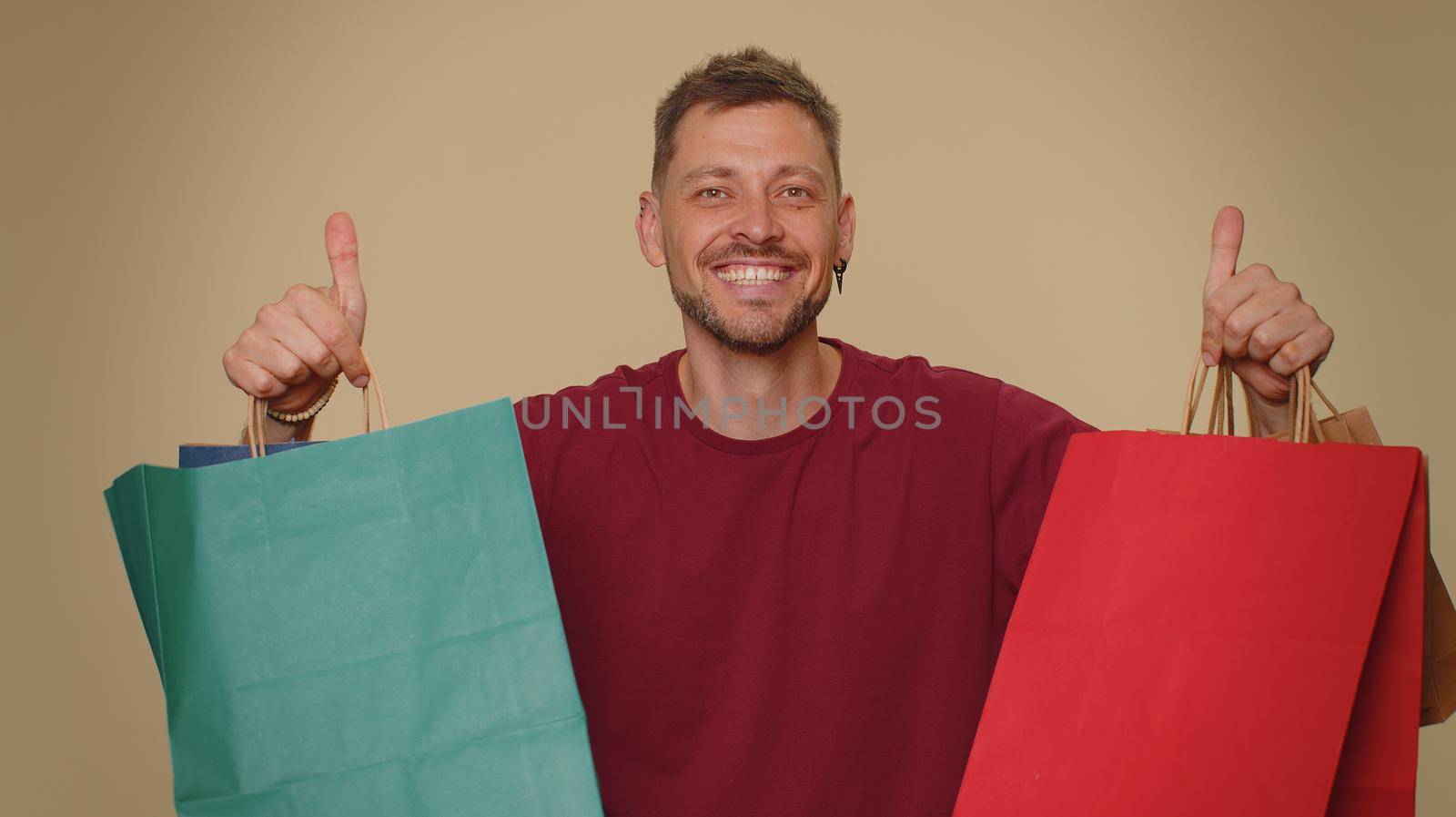 Happy young man showing shopping bags, advertising discounts, smiling looking amazed with low prices, shopping on Black Friday holidays. Adult stylish guy isolated alone on beige studio background