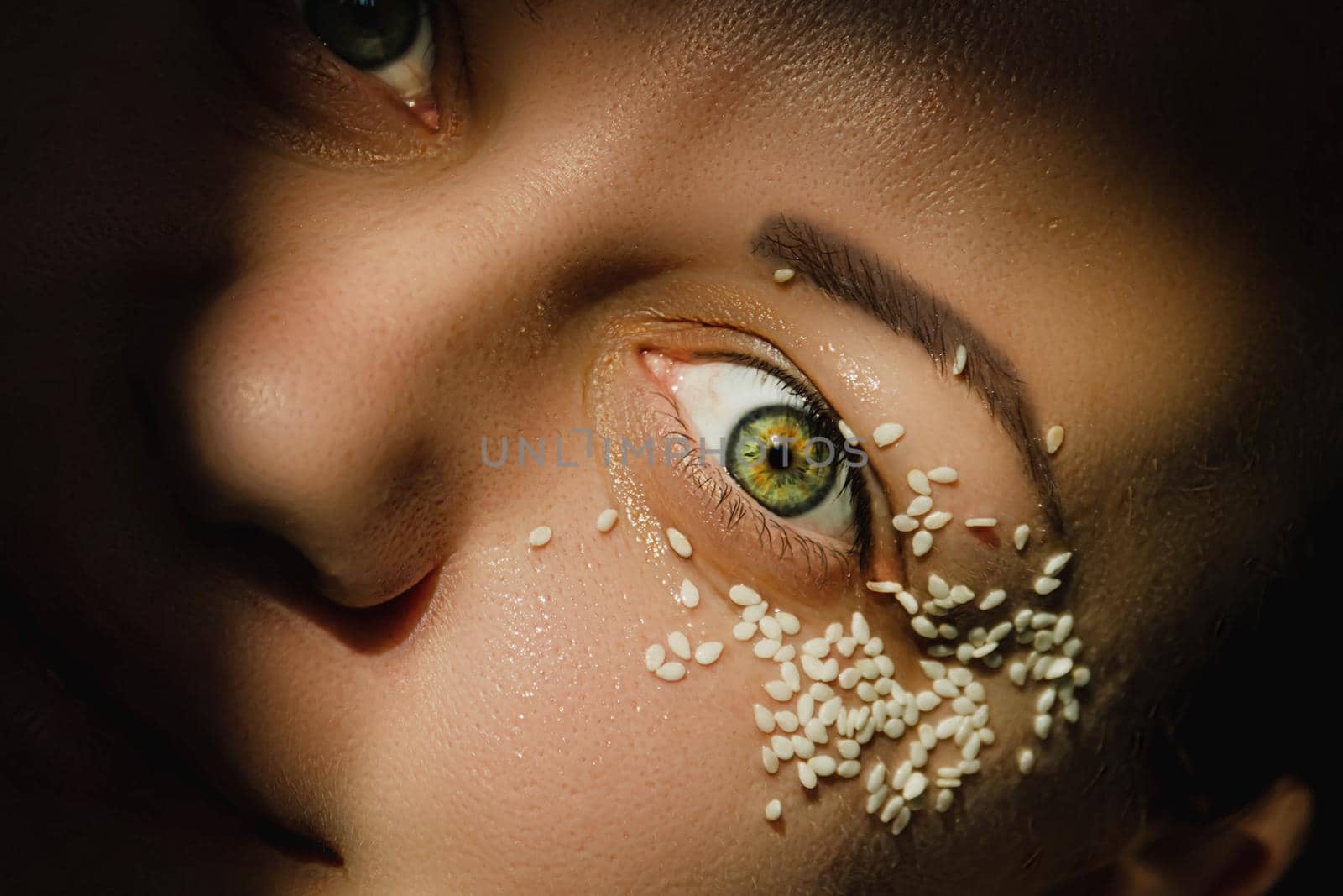 Open, green eye of a woman with white sesame seeds on the eyelid, around the eye. Expressive look