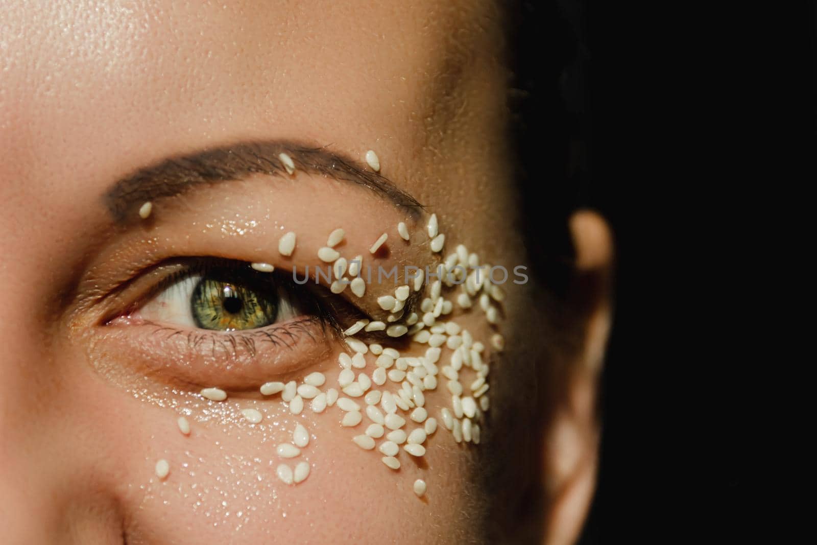 Open, green eye of a woman with white sesame seeds on the eyelid, around the eye. Expressive look