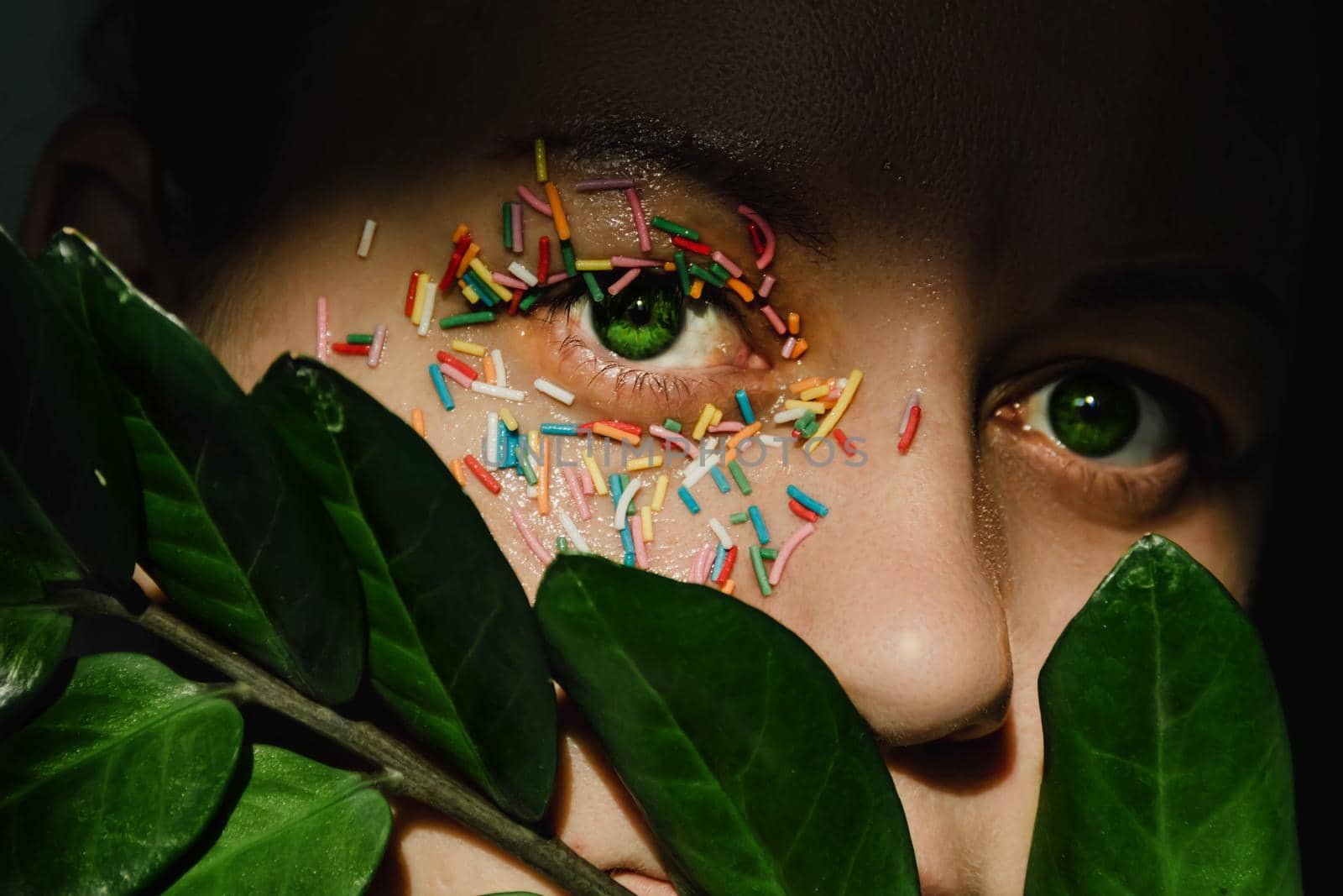 An open, green woman's eye with a sweet, multicolored sprinkle on the eyelid and a plant with large, green leaves