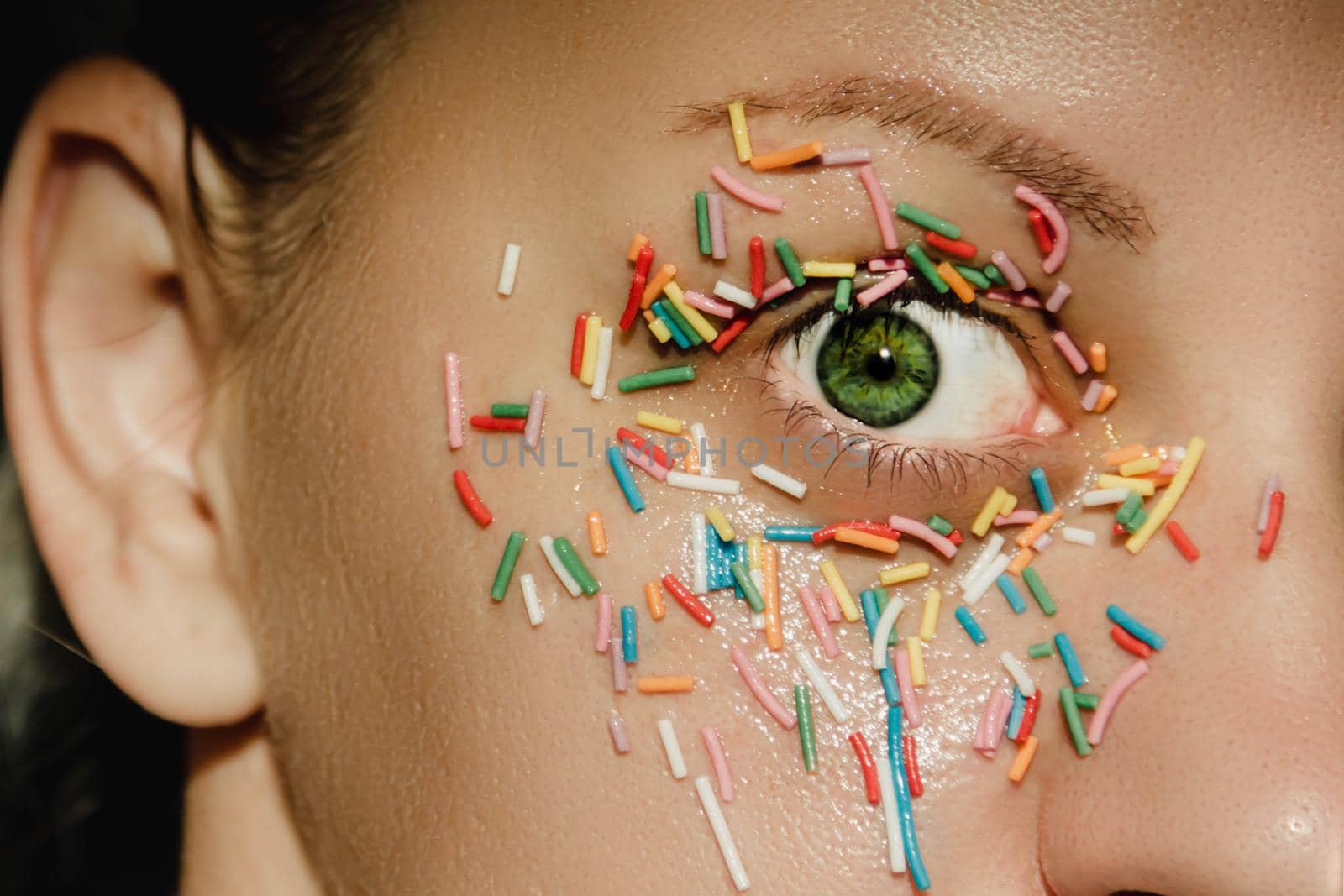 An open, green woman's eye with a sweet, multicolored sprinkle on the eyelid, around the eye