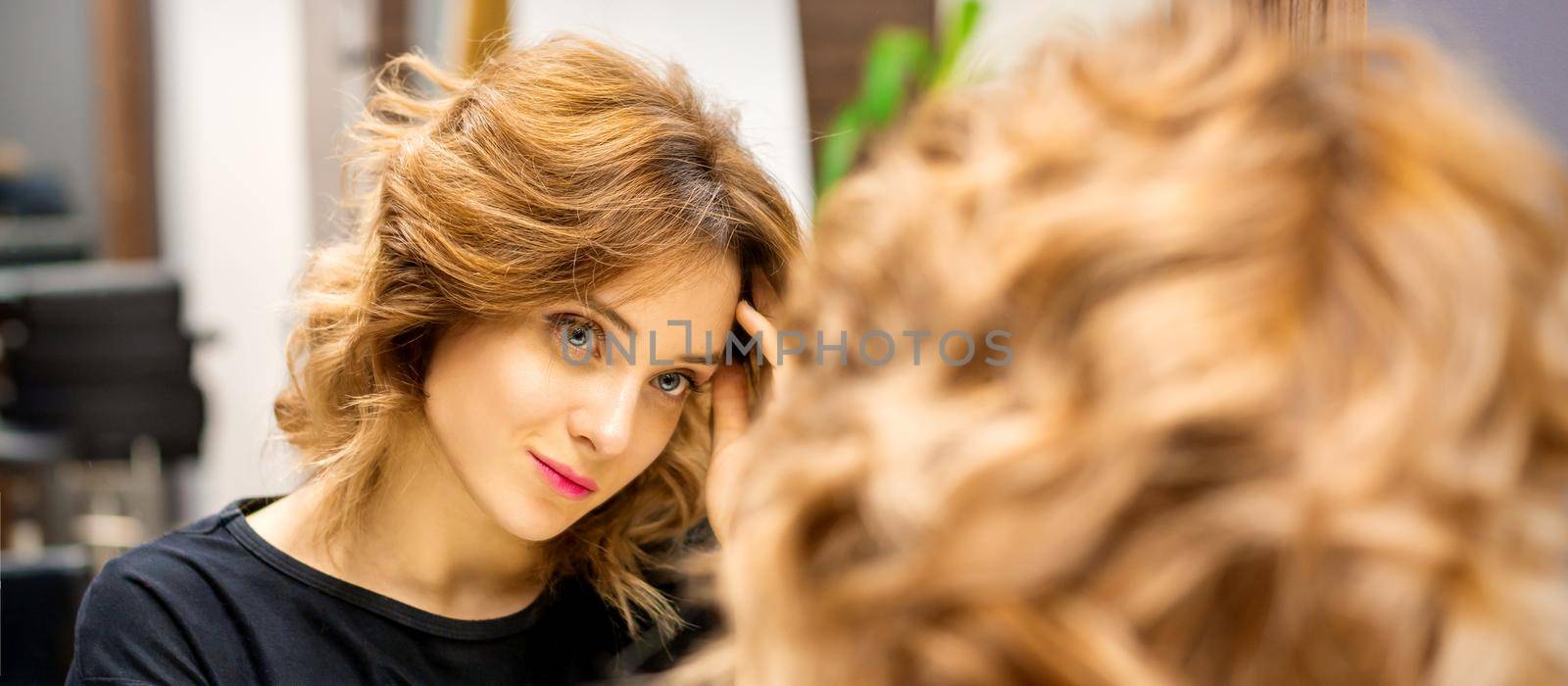 The beautiful young caucasian woman looks at her reflection in the mirror checking hairstyle and makeup in a beauty salon