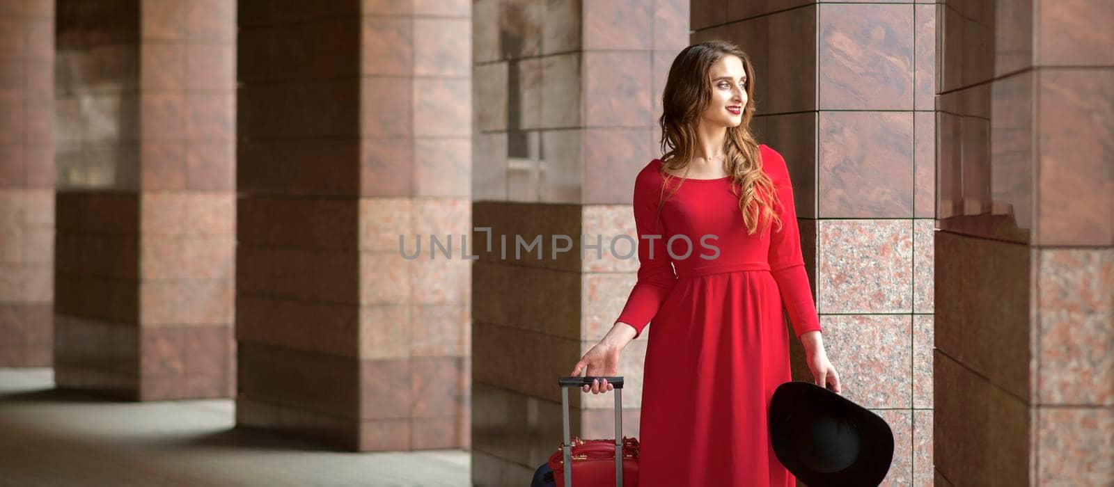 Beautiful young caucasian woman wearing red long dress with a suitcase standing in columns