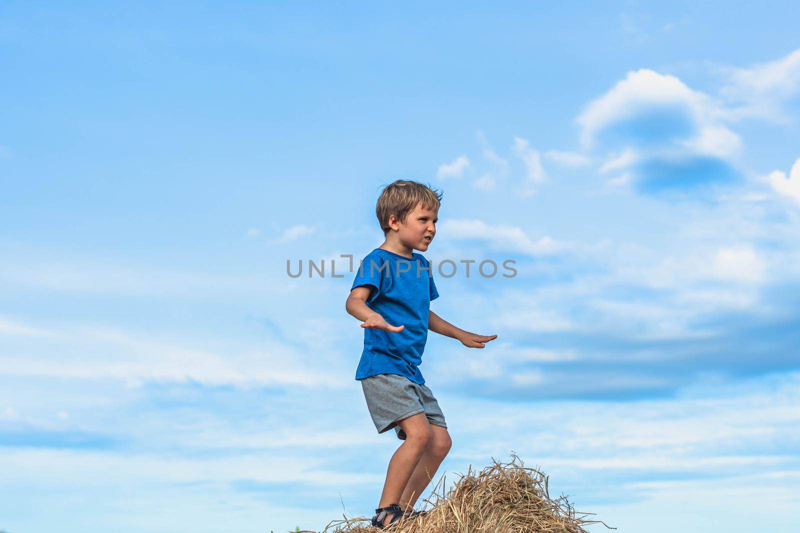 Boy smile play dance grimace show off blue t-shirt stand on haystack bales of dry grass, clear sky sunny day. Balance training. Concept happy childhood, children outdoors, clean air close to nature.