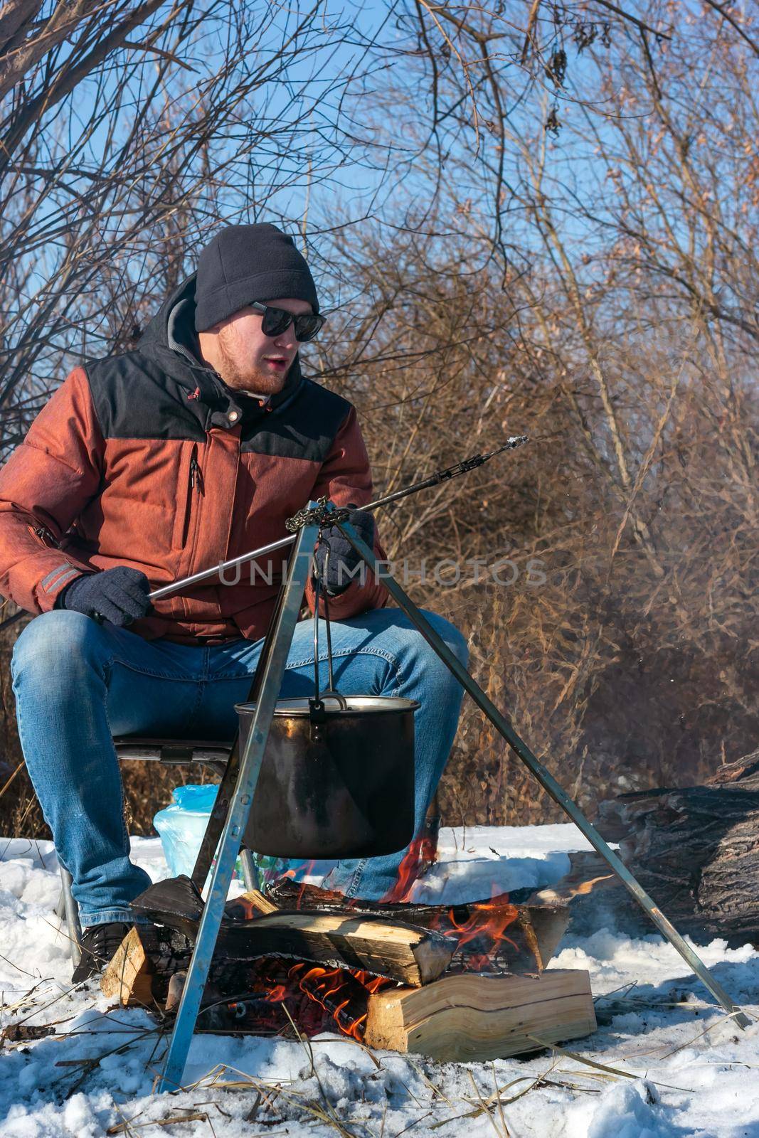 Male caucasian is sitting by campfire, pot of soot over bonfire hanging on tripod, winter outdoor cooking at campsite, lifestyle, vertikal image