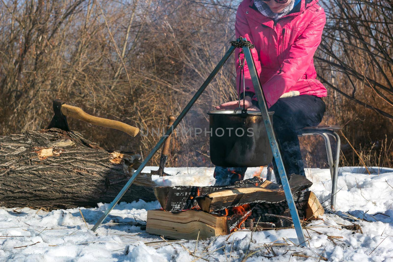 Female caucasian in pink jacket warms her hands by campfire, pot of soot over bonfire hanging on tripod, ax in tree trunk on backdrop, winter outdoor cooking at campsite, lifestyle