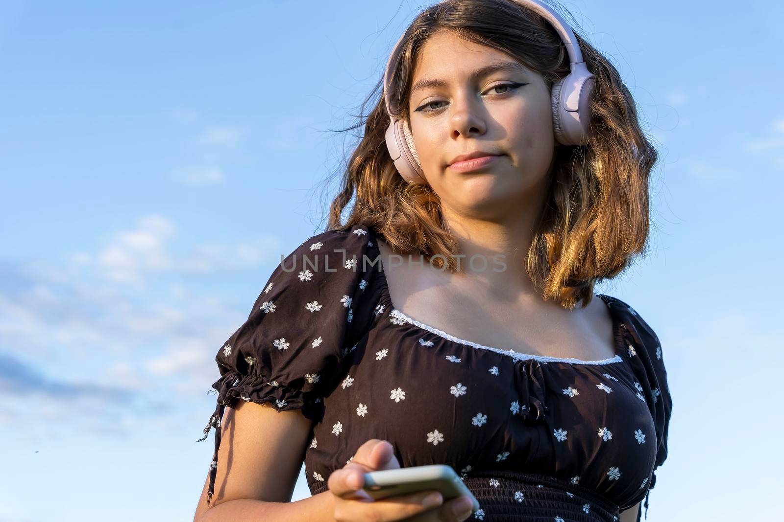 A teenage girl in a dress listens to music on her smartphone through wireless headphones against a blue sky background