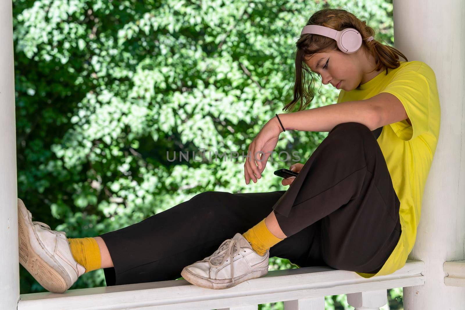 A pretty teenage girl is resting sitting on the balcony , listening to music through wireless headphones and looking at the smartphone screen