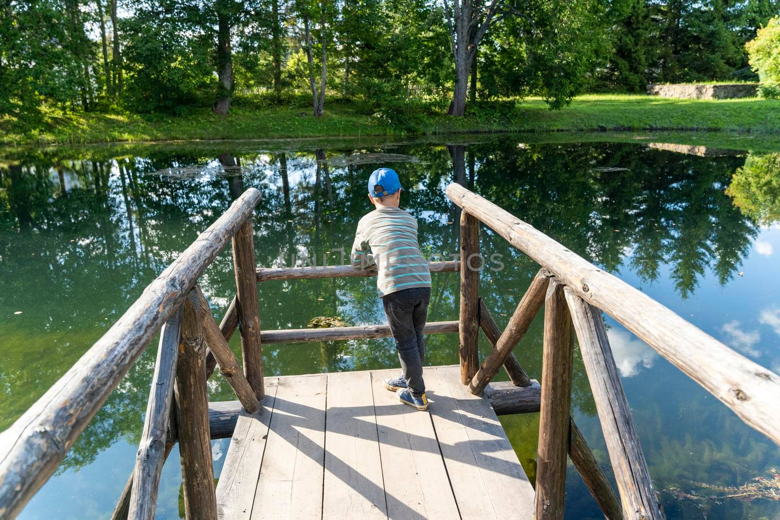 a preschooler boy is resting, leaning on the wooden bridges of the pond, walking through the nature park. A child is resting on a wooden bridge. The joys of childhood