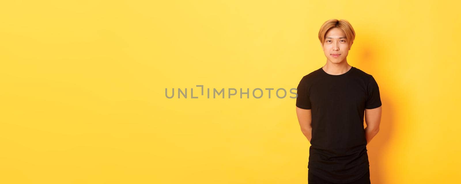 Portrait of handsome smiling asian blond guy looking sassy, holding hands behind back, standing yellow background.
