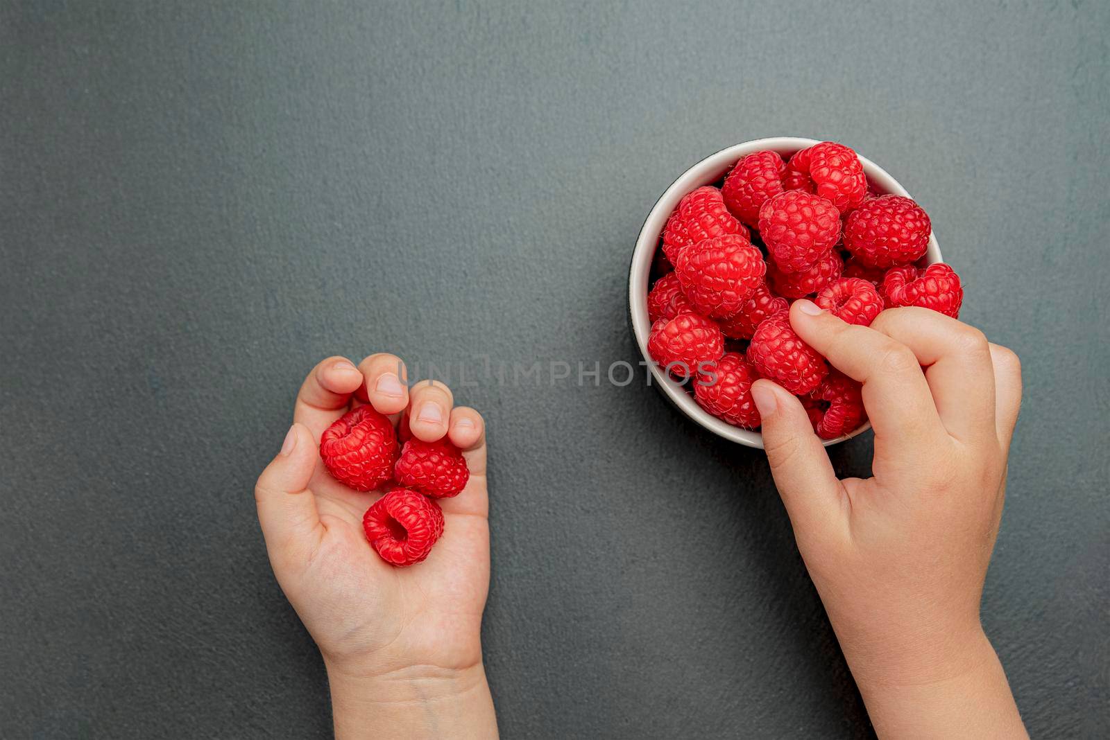 The child takes a raspberry and puts it in his hand. Berries on a black background in a plate, place to insert text, recipe or design.
