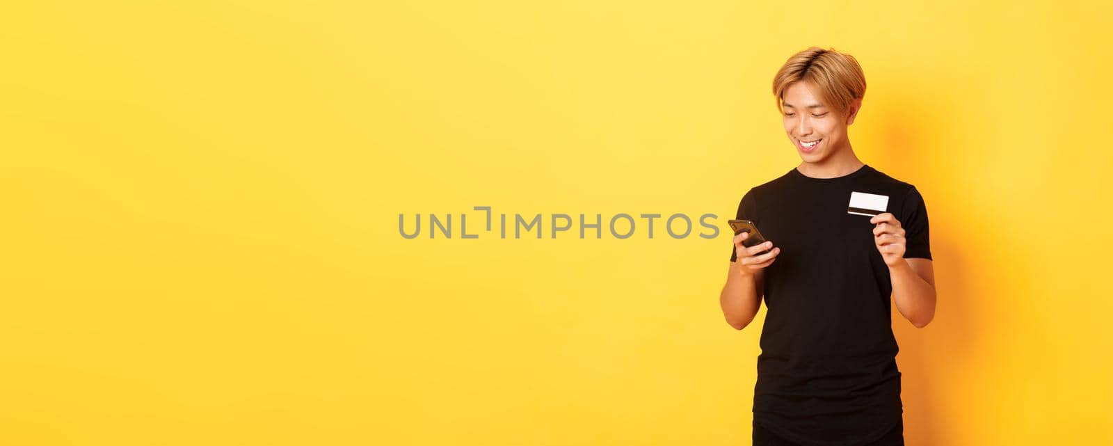 Smiling handsome asian man paying for online purchase, shopping in internet, holding smartphone and credit card, yellow background.