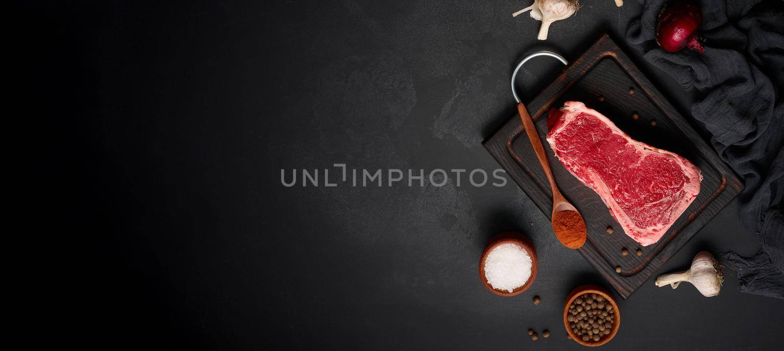Raw juicy piece of beef meat on the bone lies on a wooden cutting board, spices for cooking on a black background. Meat tenderloin New York, copy space
