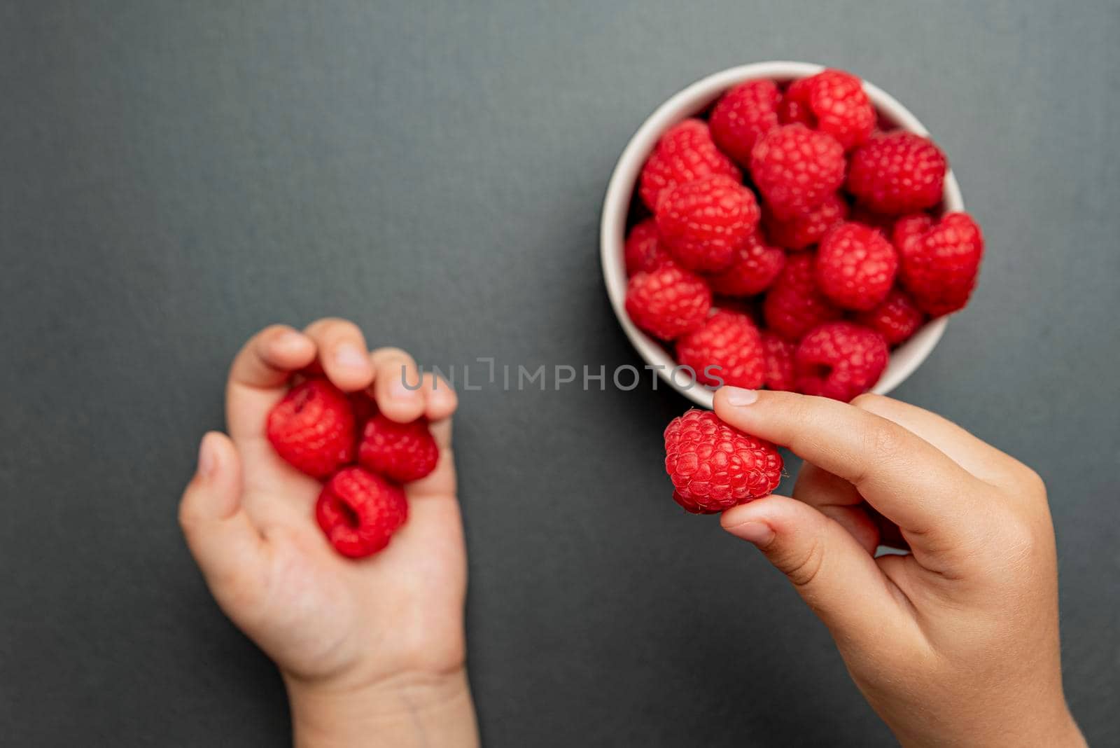 Raspberries in a saucer on a black background. The child takes raspberries from the plate and puts them in his hand. Copy space, banner on the theme of food.