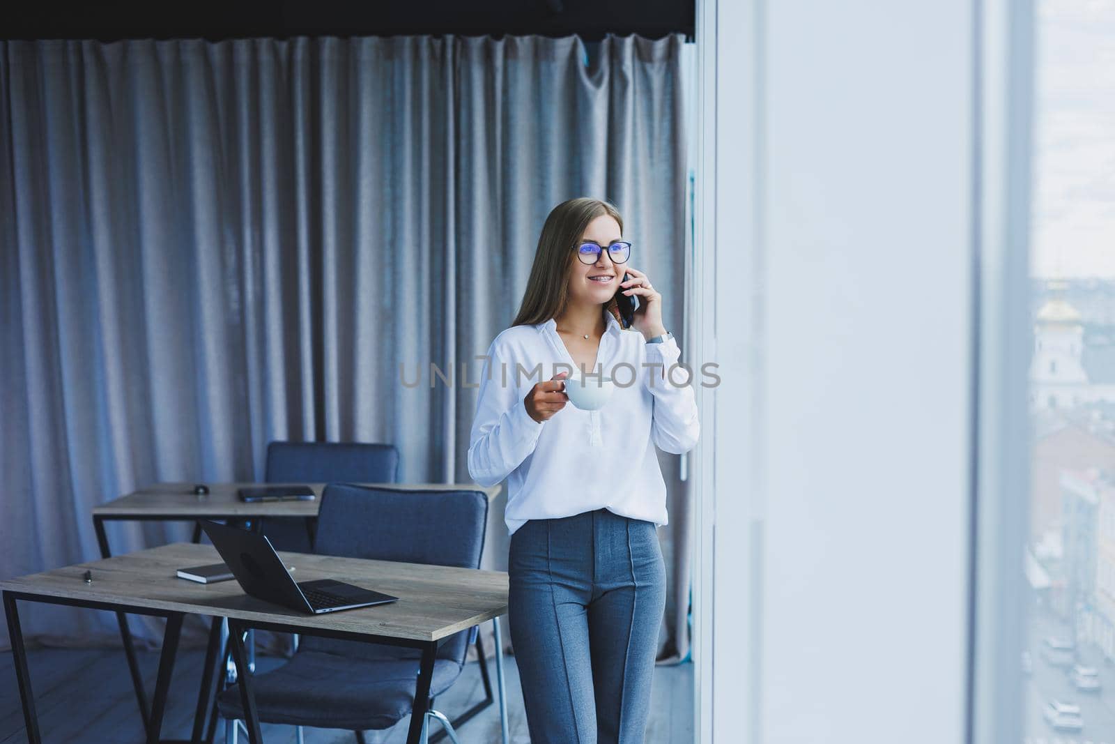 Photo of a successful young woman in a shirt smiling and working on a laptop while talking on the phone in a modern office with large windows. Remote work with coffee