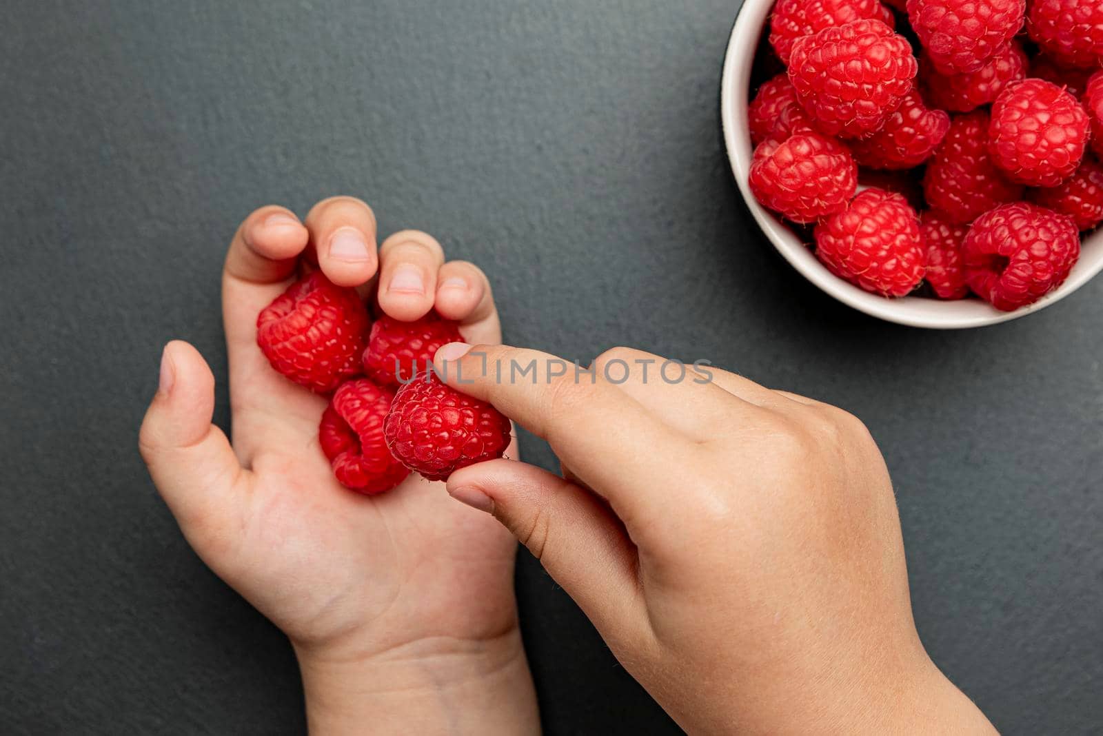 Raspberries in a saucer on a black background. The child takes raspberries from the plate and puts them in his hand. Copy space, banner on the theme of food.