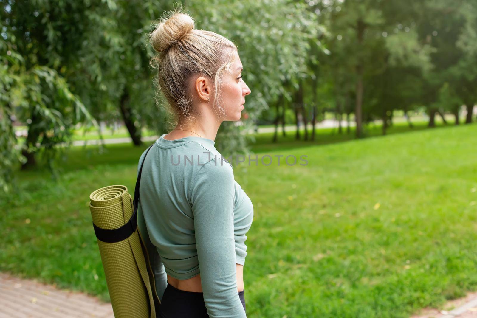 A blonde woman in a green top, standing in summer, in park, with a green gym mat for sports. side view. Close up. Copy space