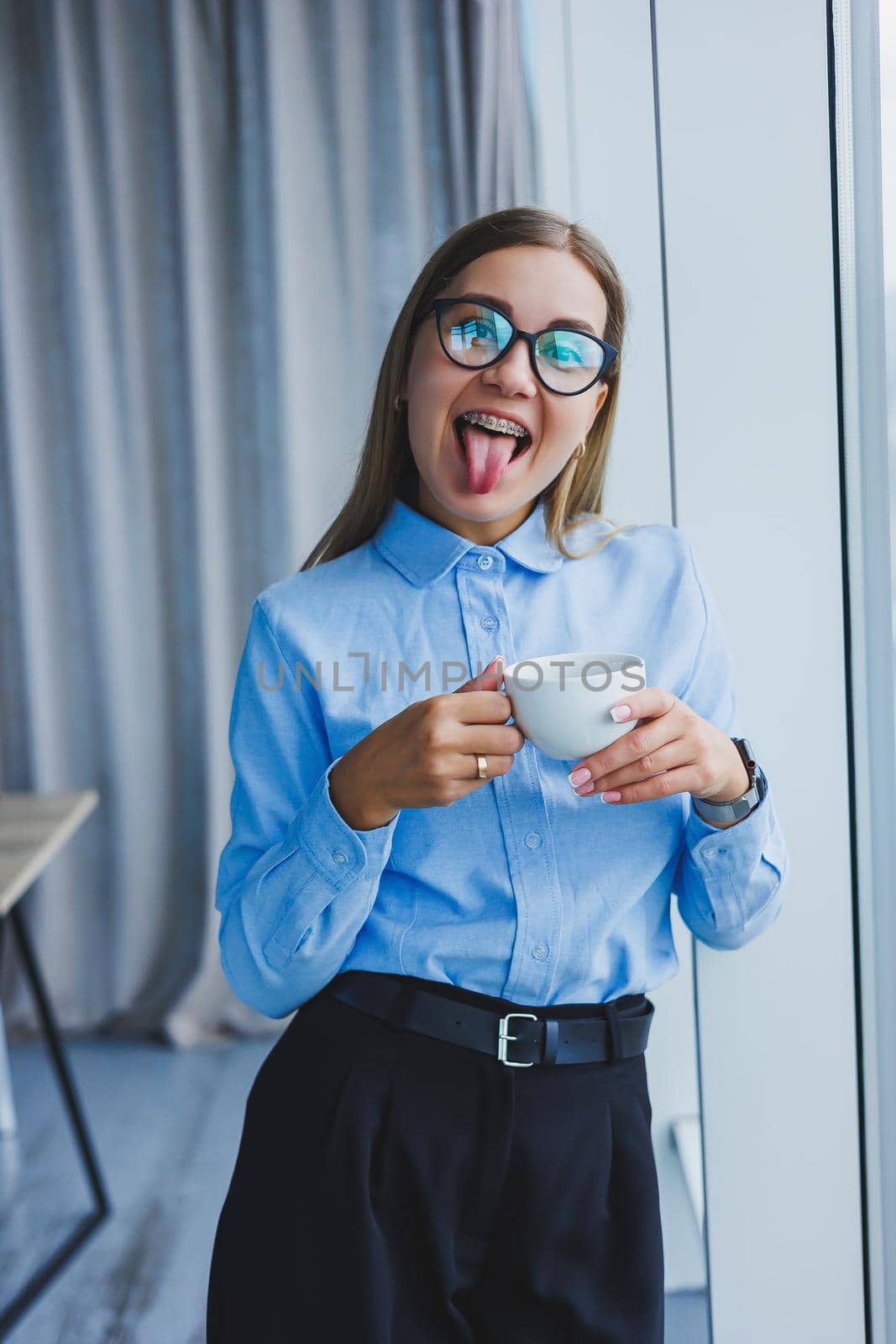 Image of a happy young woman in a classic shirt and glasses smiling and drinking coffee while standing by the window in a modern office with large windows. Remote work