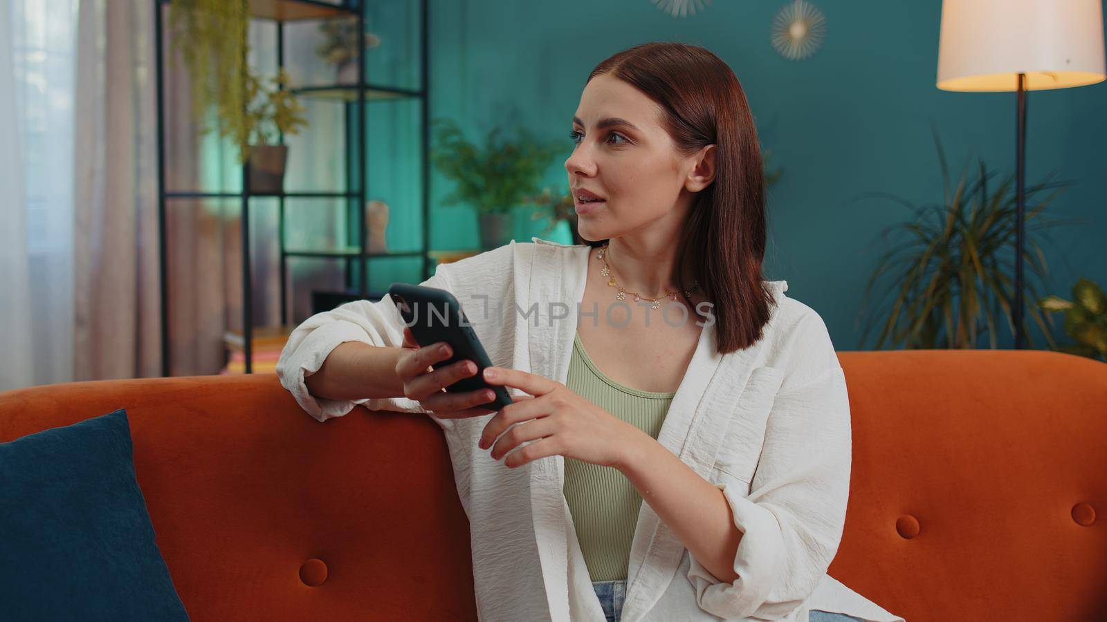 Portrait of adult girl sitting on sofa uses mobile phone smile at modern home apartment. Young woman texting share messages content on smartphone social media applications online, watching relax movie