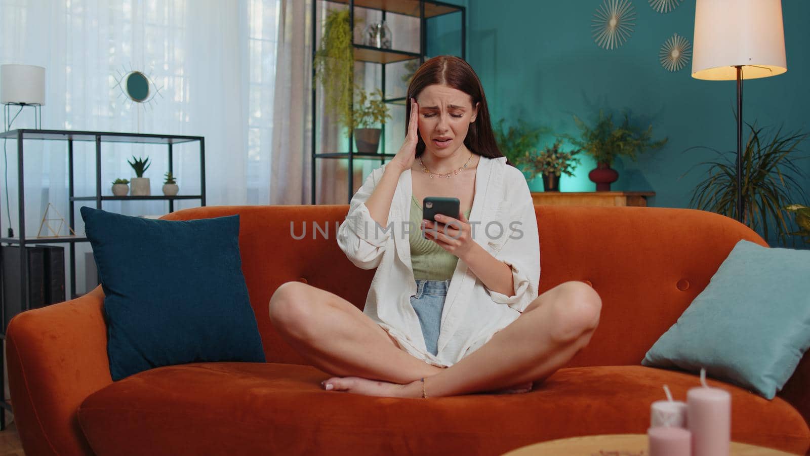 Portrait of caucasian adult girl use smartphone typing browsing, loses becoming surprised sudden lottery results, bad news, fortune loss, fail. Young woman sitting on orange couch at home living room