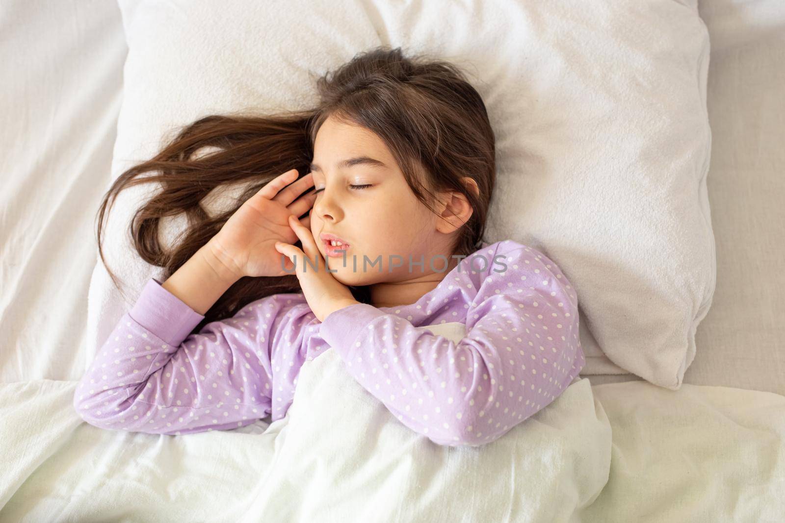 Little charming brunette girl in purple pajamas, sleeps lying on a white pillow on the bed. View from above. Top view. Copy space