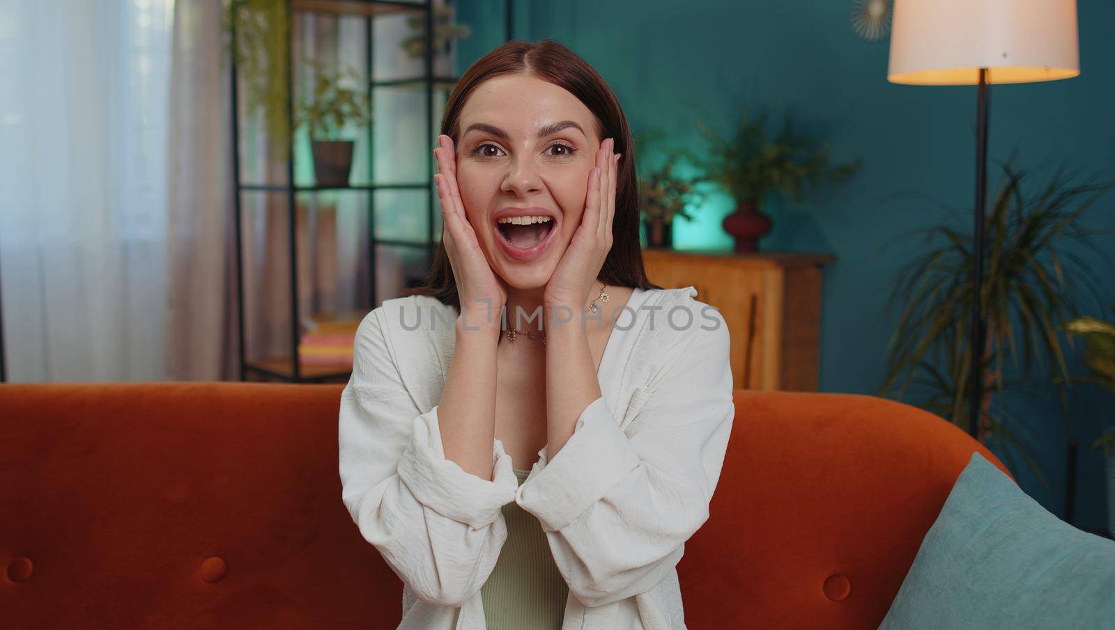Oh my God, Wow. Lovely adult girl surprised looking at camera with big eyes, shocked by sudden victory, good win news, celebrating at home. Excited amazed young woman indoors in living room on couch