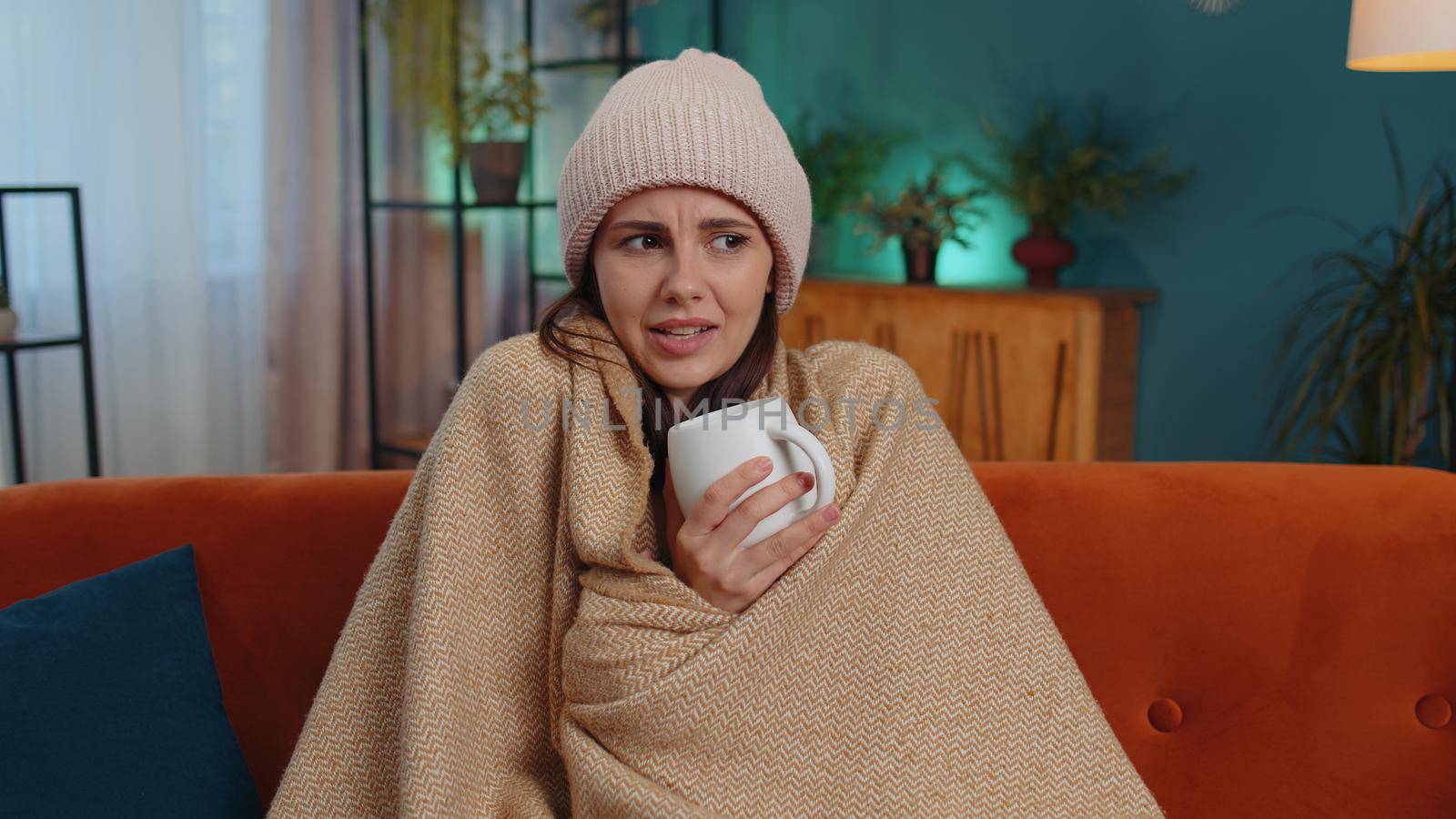 Sick girl wear hat wrapped in plaid sit alone shivering from cold on sofa drinking hot tea in unheated apartment without heating due debt. Unhealthy young woman feeling discomfort try to warming up