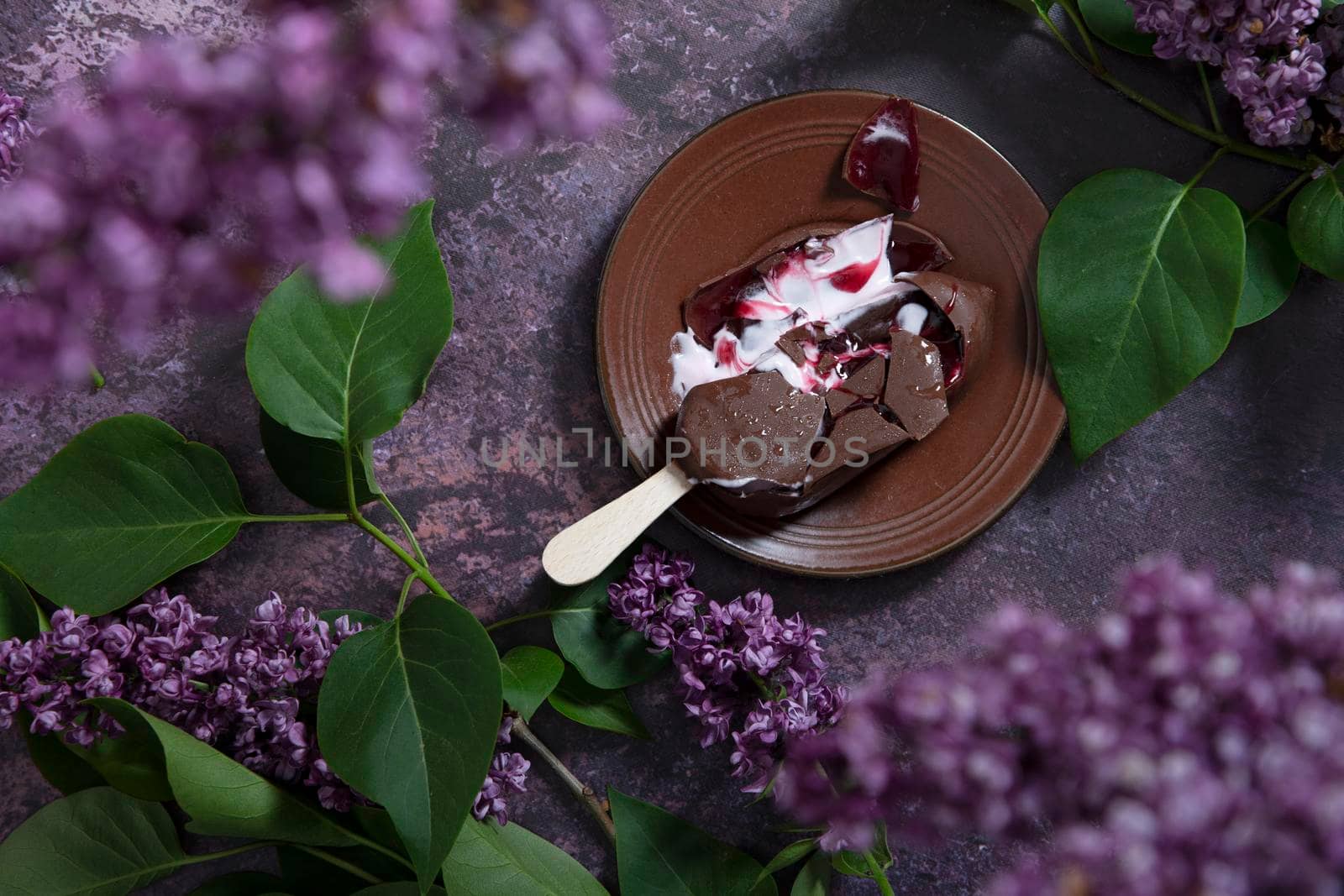 melted raspberry ice cream in chocolate on a wooden stick on a dark burgundy background with lilac flowers, a sweet dessert on a hot spring day, flat lay food. High quality photo