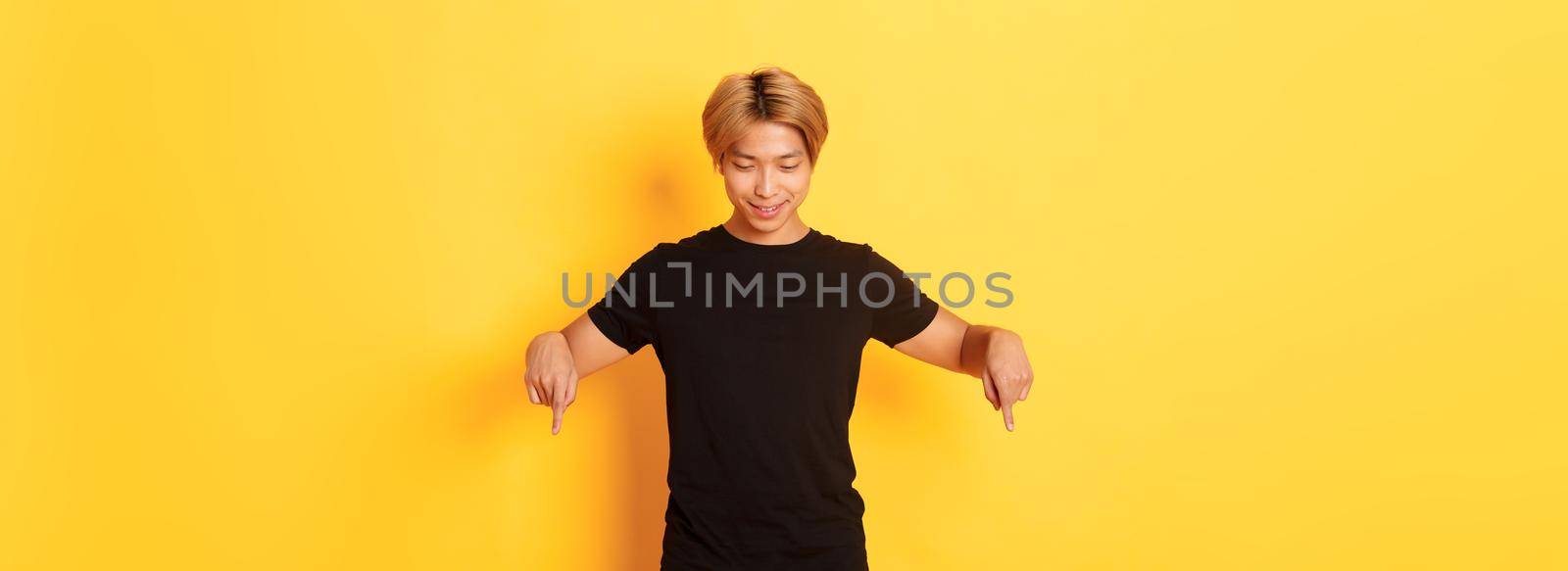 Portrait of curious happy blond asian guy, looking and pointing fingers down with pleased smile, yellow background.