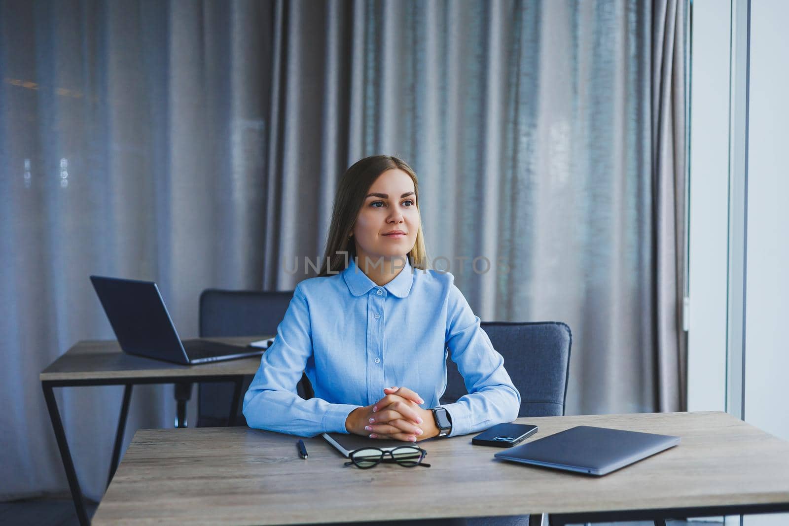 A beautiful woman in glasses sits at a table in the office and works at a laptop. Corporate work in the office. Modern woman workplace manager in classic clothes