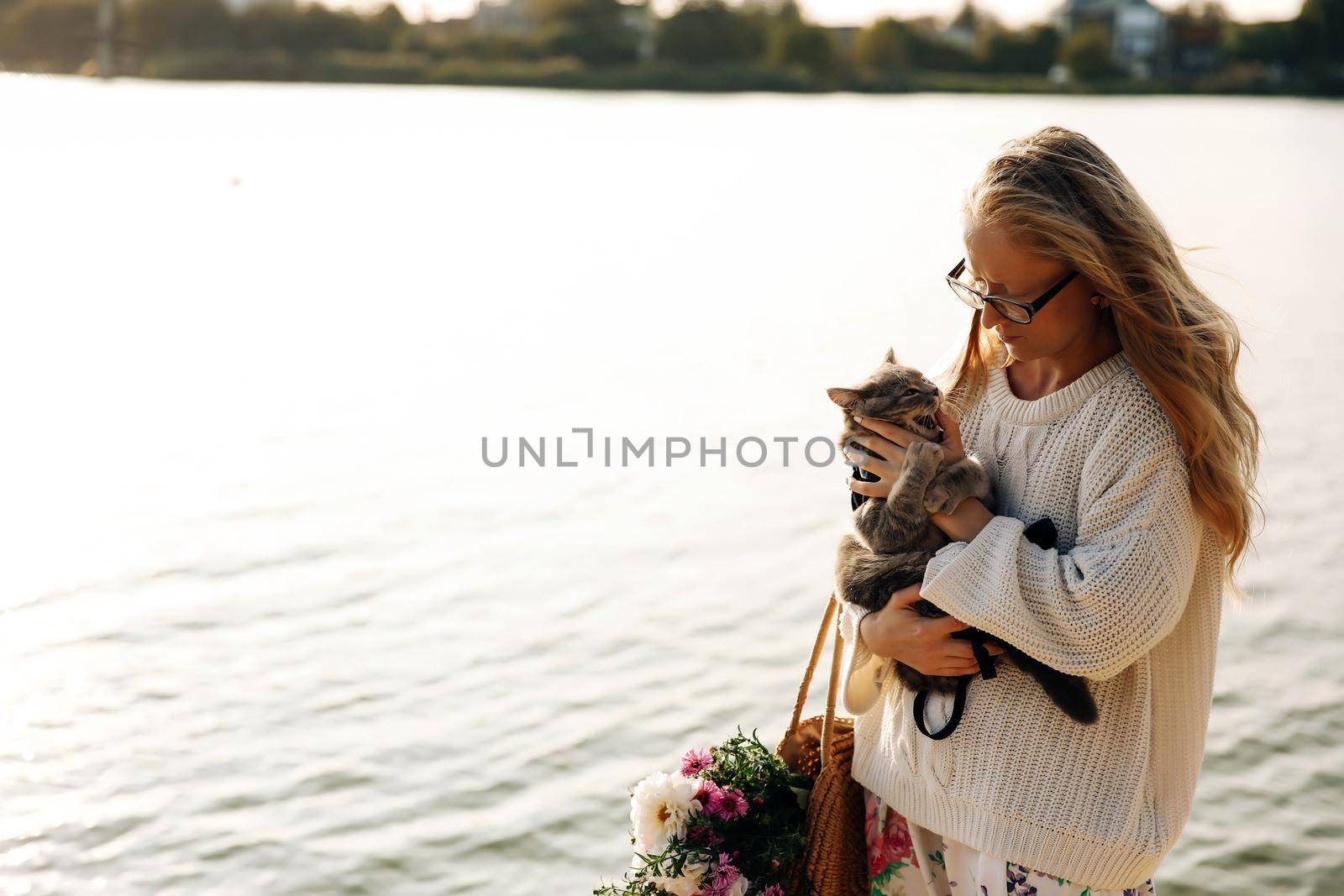 Young blonde, girl, woman with glasses holds a cat breed Scottish Straight, the cat bites. Walk with pet near the lake, river at sunset on the beach in Spring
