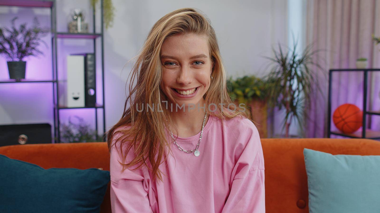 Close-up portrait of happy smiling caucasian girl in hoodie, looking at camera, celebrate good news. Young woman indoor isolated at home in living room sitting on orange couch. Female nature beauty