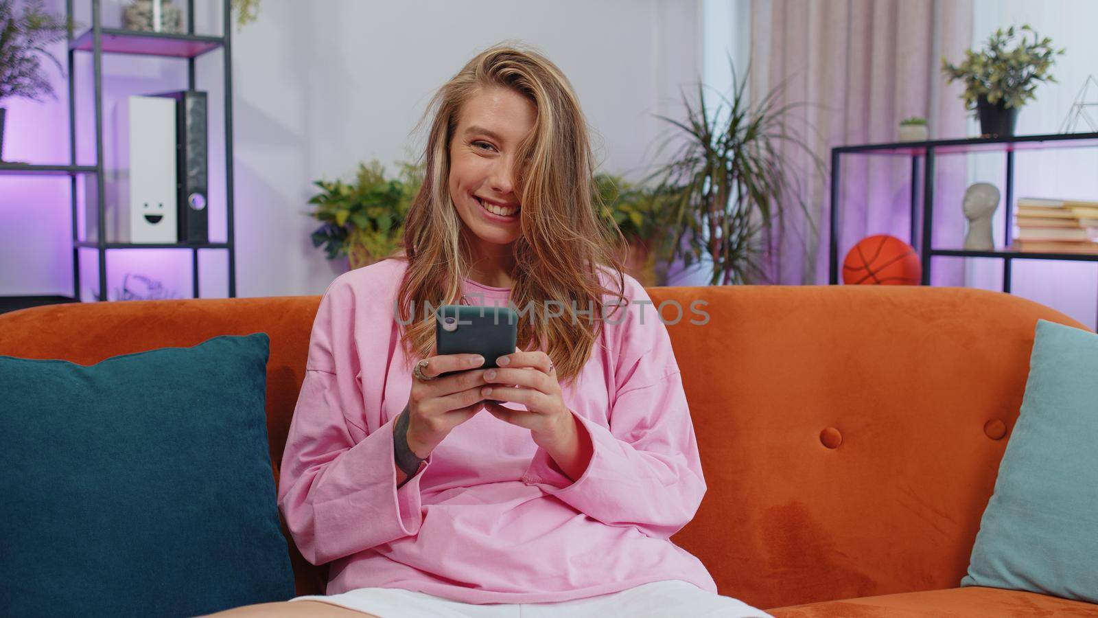Portrait of adult girl sitting on sofa uses mobile phone smile at modern home apartment. Young woman texting share messages content on smartphone social media applications online, watching relax movie