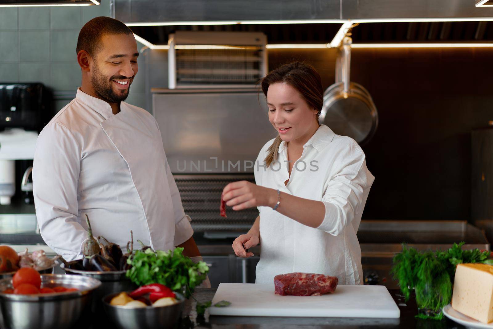 Handsome young African chef is cooking together with Caucasian girlfriend in the kitchen A cook teaches a girl how to cook. Man and woman cooking in professional kitchen. interracial relationship