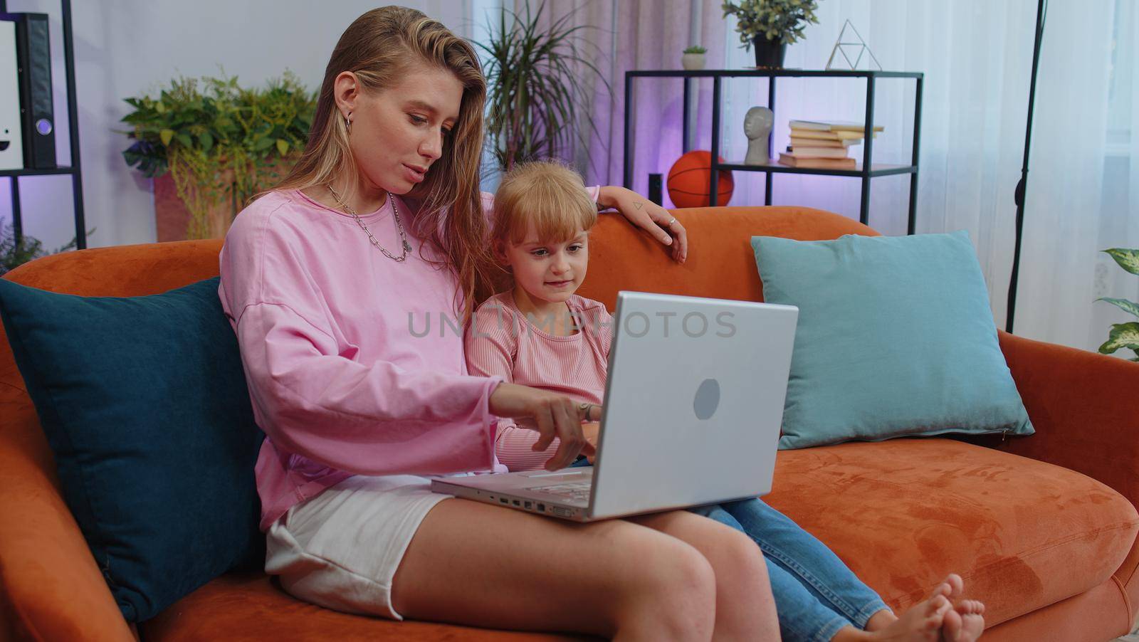 Happy family mother or nanny and child kid daughter laugh use laptop, watch cartoons, make video call, look at screen. Mom with child making online shopping on notebook sit on sofa at home living room