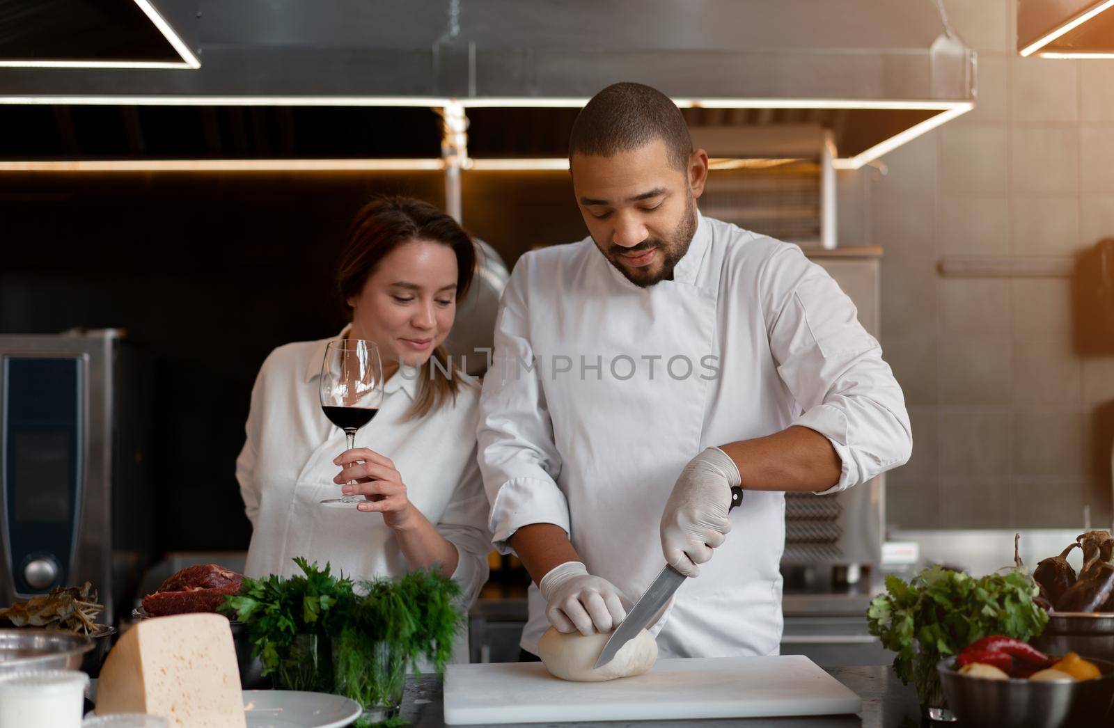 Handsome young African chef is cooking together with his Caucasian girlfriend in the kitchen using red wine as an ingredient. by andreonegin