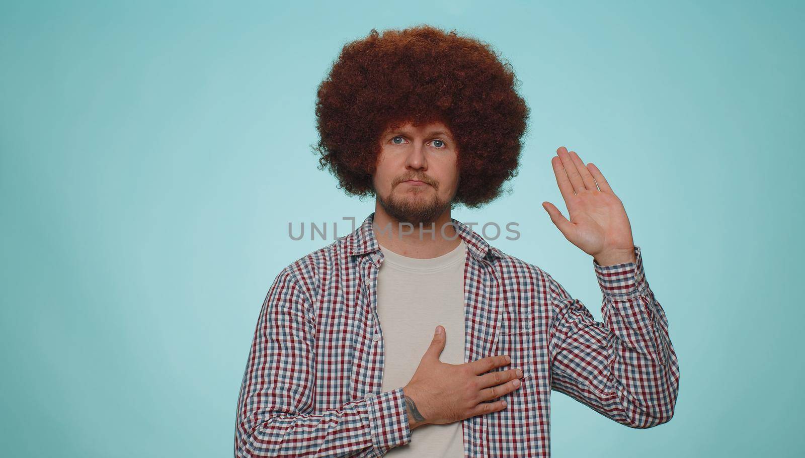 I swear to be honest. Sincere responsible man raising hand to take oath, promising to be honest and to tell truth, keeping hand on chest. Young adult guy boy isolated alone on blue studio background