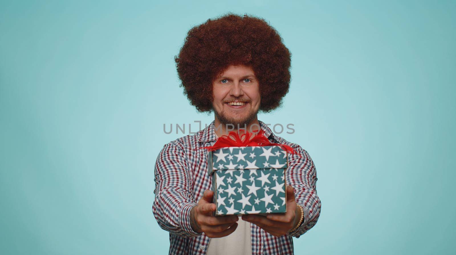 Positive smiling bearded man presenting birthday gift box stretches out hands, offer wrapped present career bonus, celebrating party. Young adult guy boy isolated alone on blue studio wall background