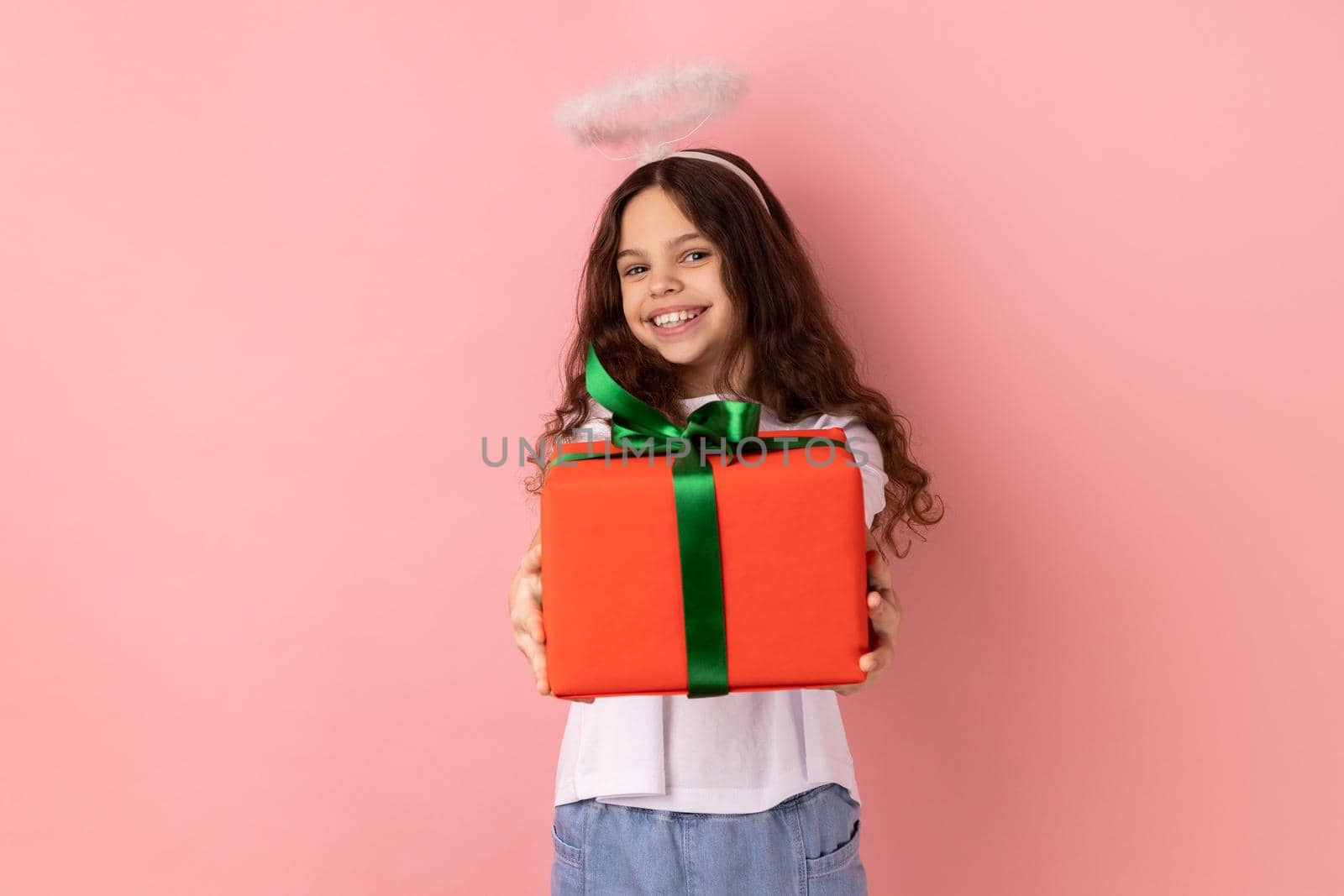 Portrait of joyful cheerful little girl wearing white T-shirt and nimb over head, holding out wrapped present box, congratulating with holiday. Indoor studio shot isolated on pink background.
